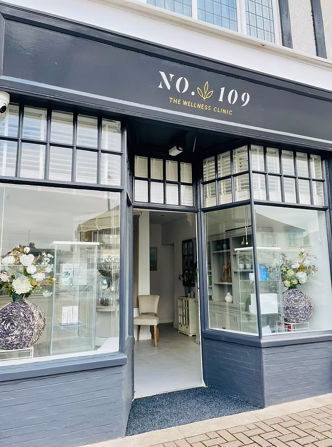 Exterior view of N° 109 The Wellness Clinic with large glass windows and a door, floral arrangements in vases inside the window displays, and a small mat outside the entrance.