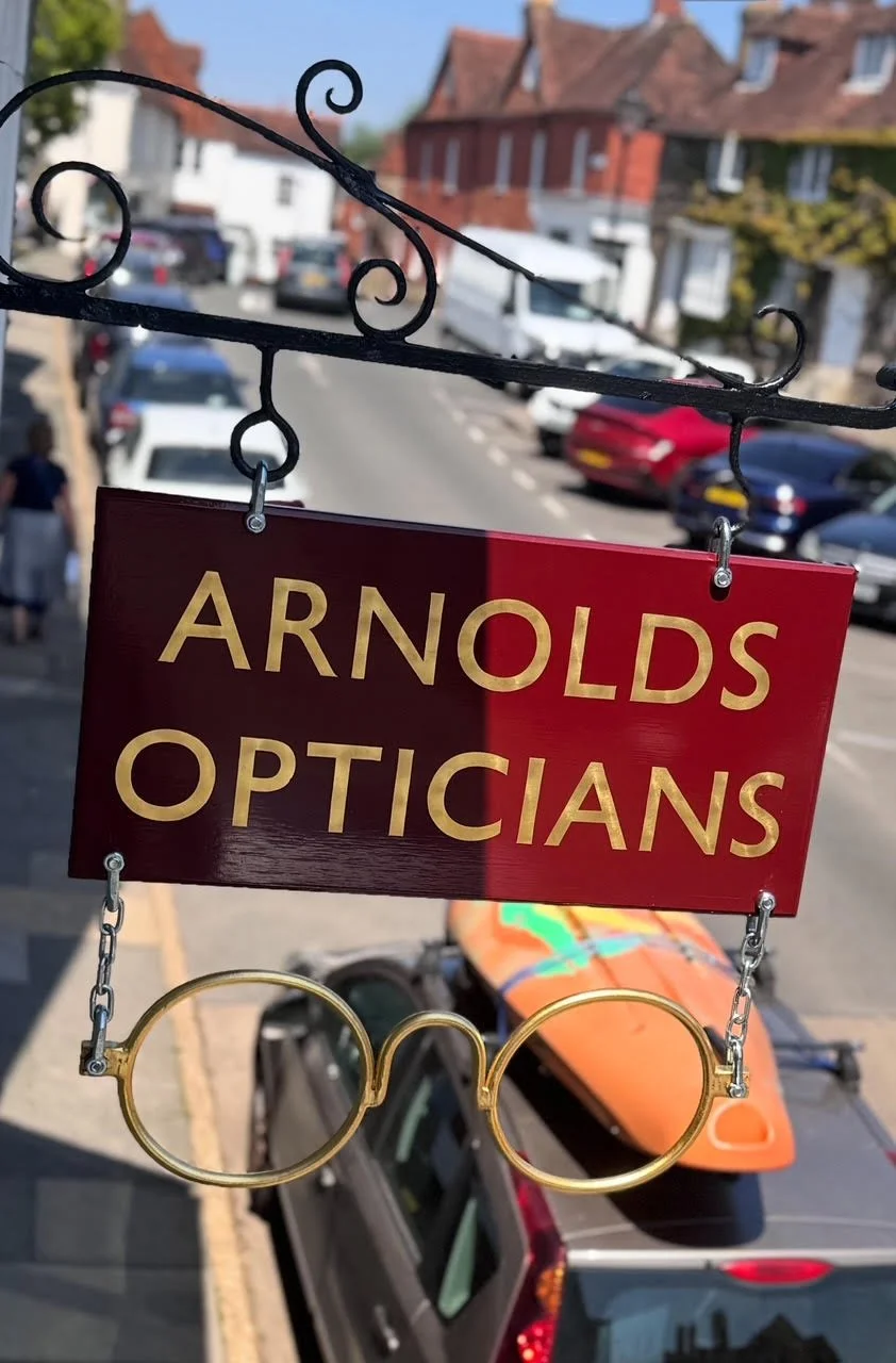 Sign for 'Arnolds Opticians' hanging outside in a neighborhood street with parked cars and houses in the background.