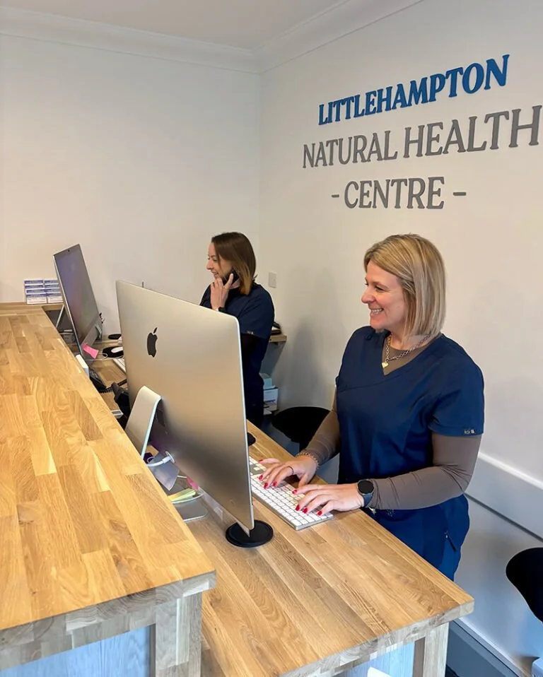 Two women in medical scrubs working at a reception desk inside a health center, with a sign on the wall reading 'Littlehampton Natural Health Centre.'
