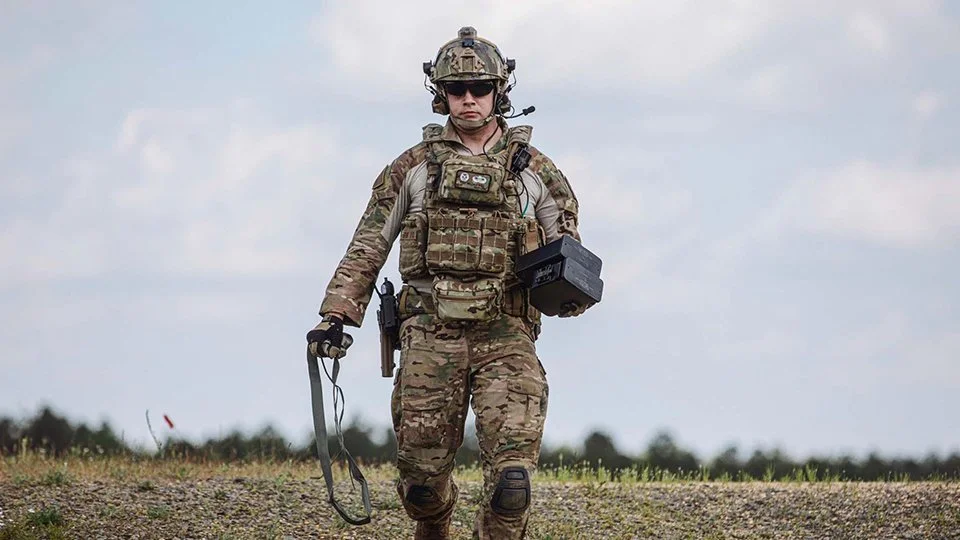 Militärperson in Camouflage auf einer offenen Wiese, mit Helm, Sonnenbrille und Ausrüstung, hält ein Kabel in der Hand und trägt eine schwarze Kiste.