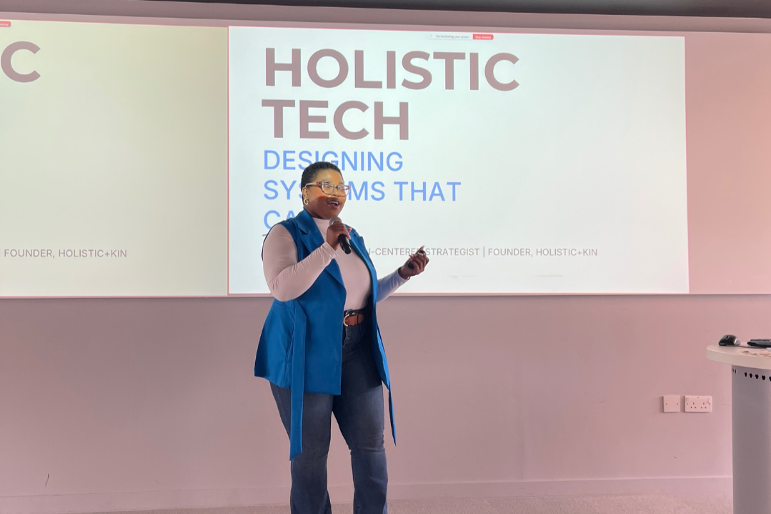 Rai Gethers giving a presentation on holistic tech and designing systems, standing in front of a large slide titled 'HOLISTIC TECH' with subtitle 'DESIGNING SYSTEMS THAT CARE. She's wearing glasses, a white long-sleeve shirt, & a blue sleeveless vest