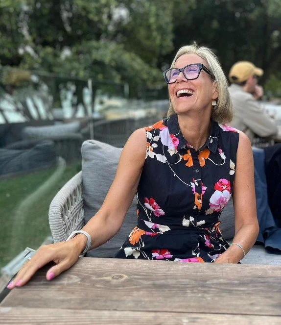 A smiling woman with glasses and short gray hair, wearing a sleeveless floral dress, sitting outdoors at a wooden table.