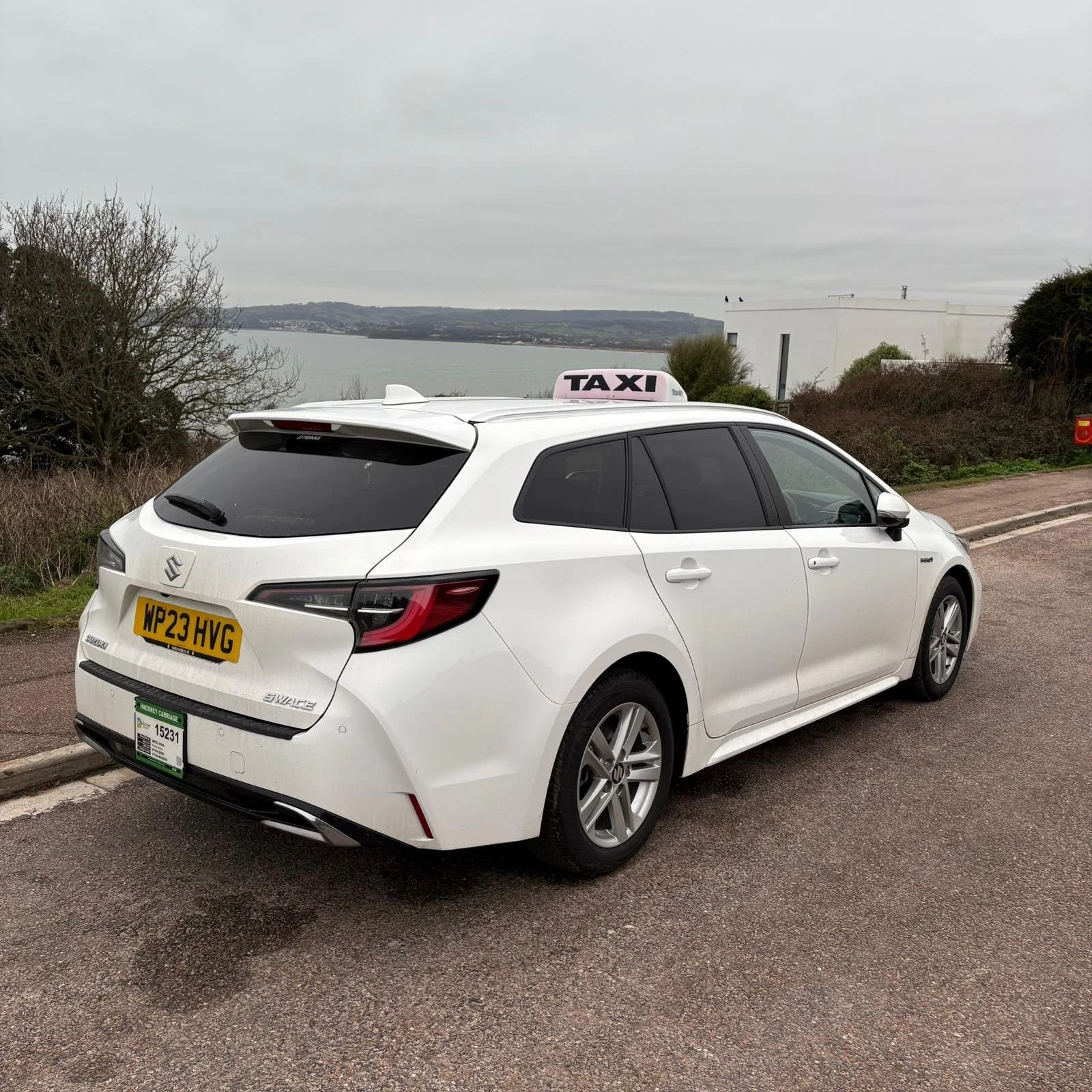 A black taxi car with 'Jurassic Taxis' written on it, parked on a paved road with a scenic ocean view, rocky cliffs, and a stone house in the background under a partly cloudy sky.