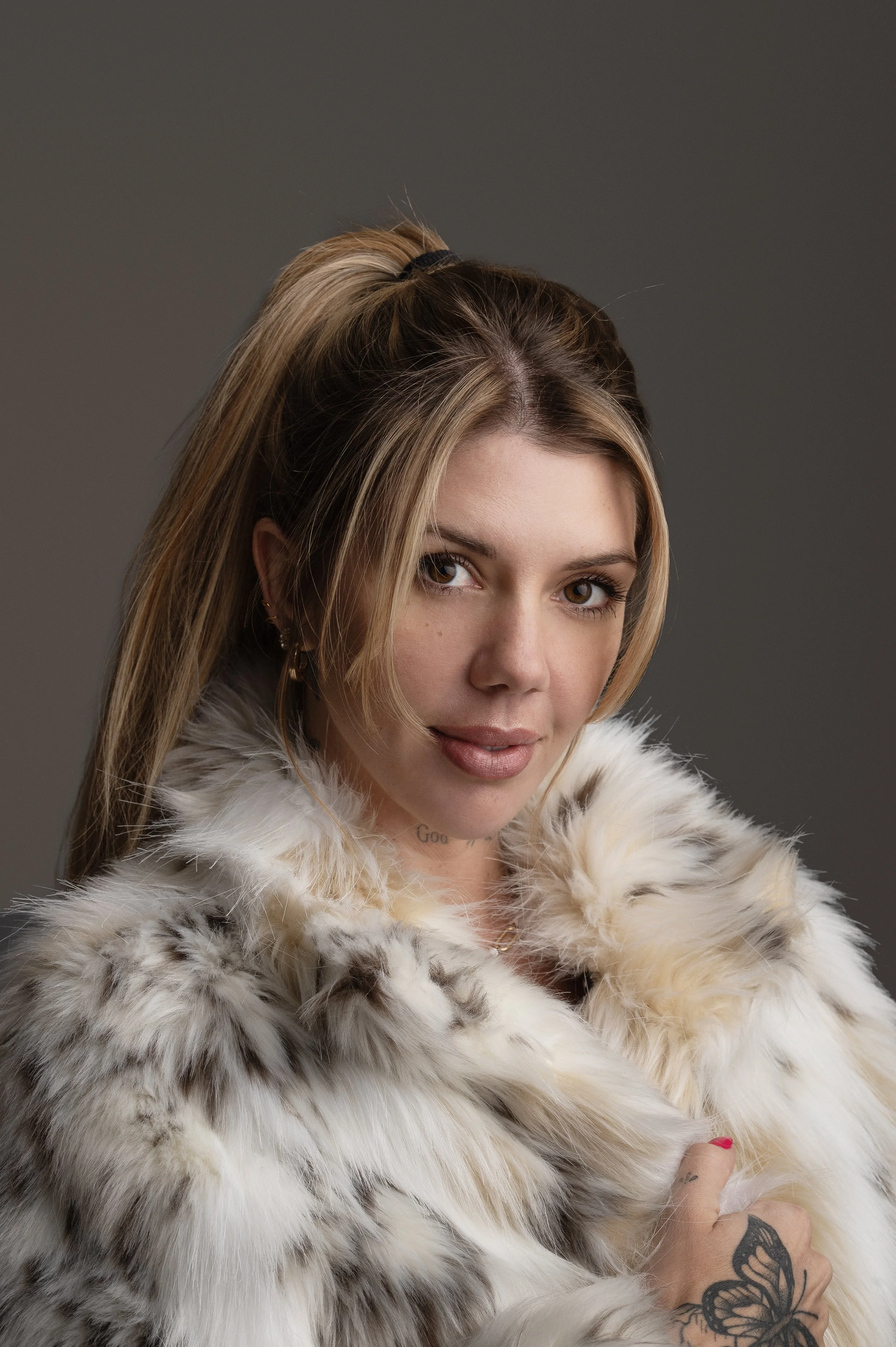 Close-up portrait of a woman with long blonde hair tied in a ponytail, wearing a white and gray fur coat, against a gray background.