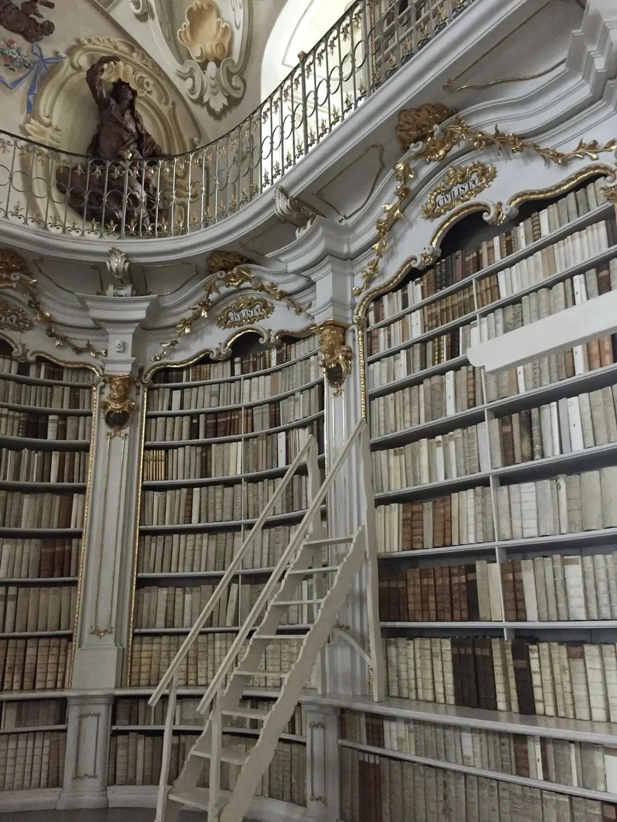 A grand, ornate library with tall bookshelves filled with old books, a white staircase, and a decorative railing. There is a sculpture of a woman on the upper level near the ceiling.
