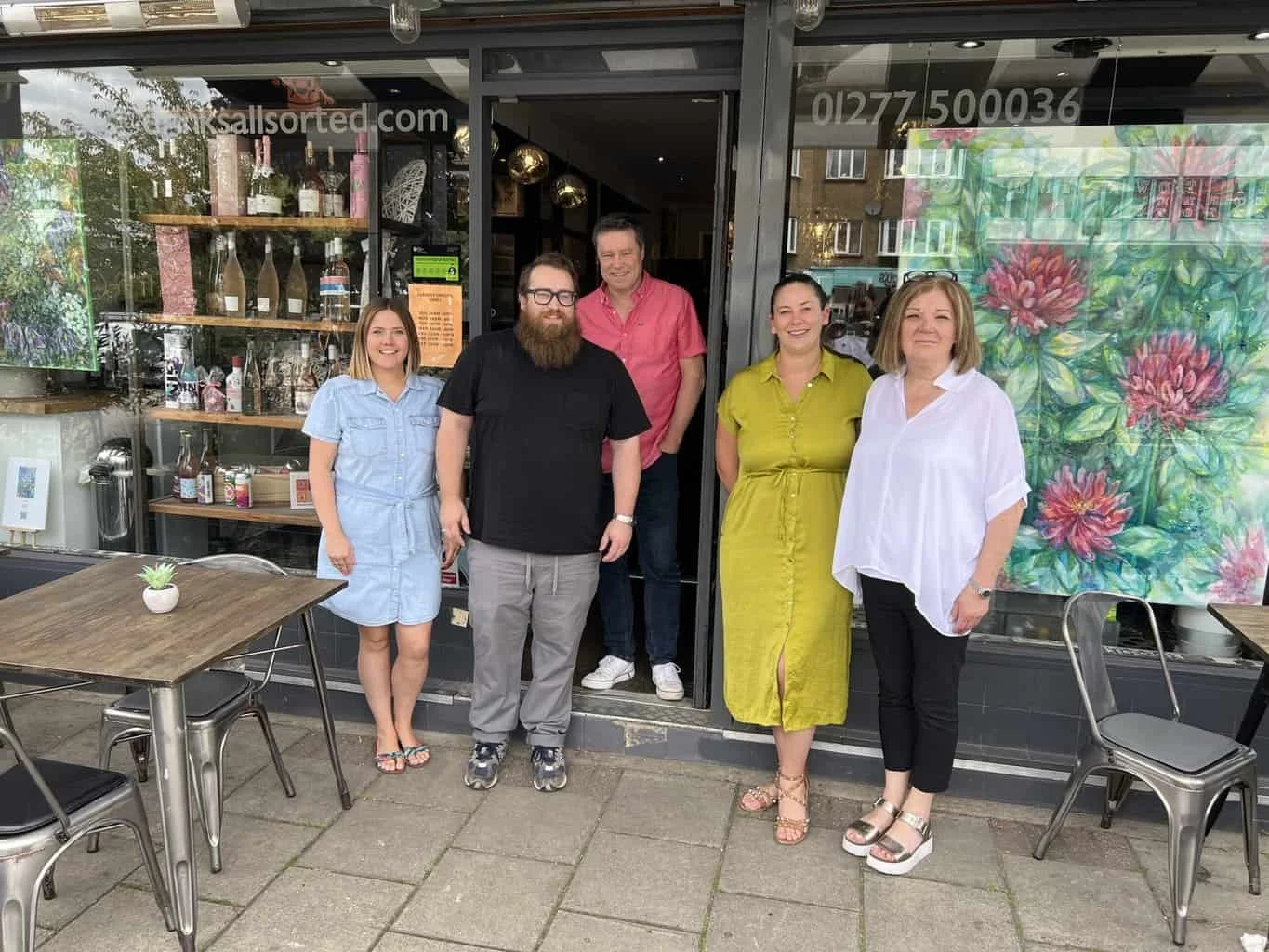Five people standing in front of a cafe, posing for a photo. There are tables and chairs outside, with one small potted plant on a table. The cafe window displays decorations and artwork, with a large colorful painting on the right side.