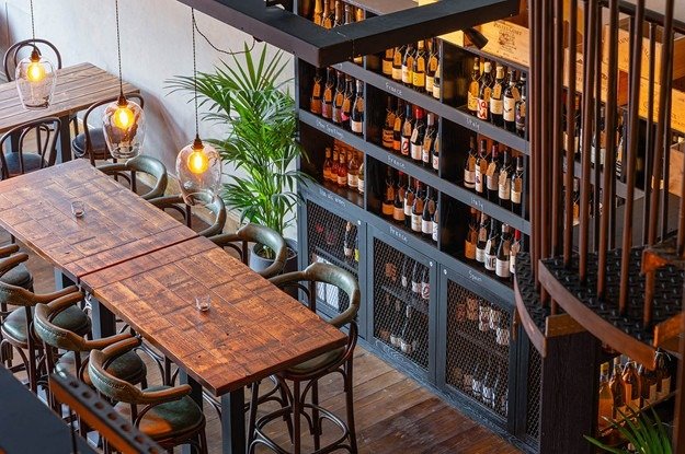 Interior of a restaurant or bar with a long wooden table, green chairs, hanging pendant lights, a large green plant, and a wine display shelf with bottles stored on racks.