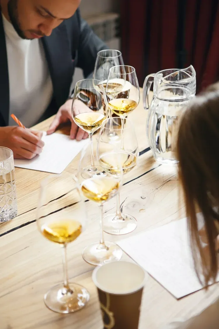 A person taking notes at a tasting event with several glasses of white wine and a water pitcher on a wooden table.