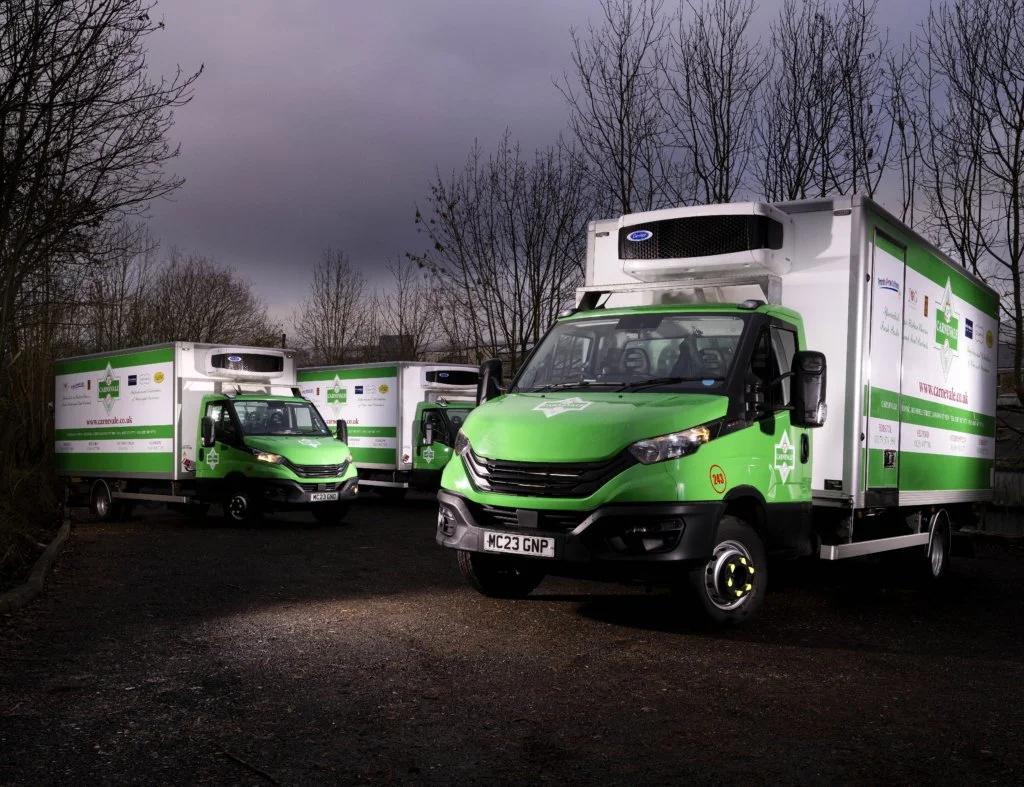 Three green and white delivery trucks parked on a dark, cloudy day with leafless trees in the background.