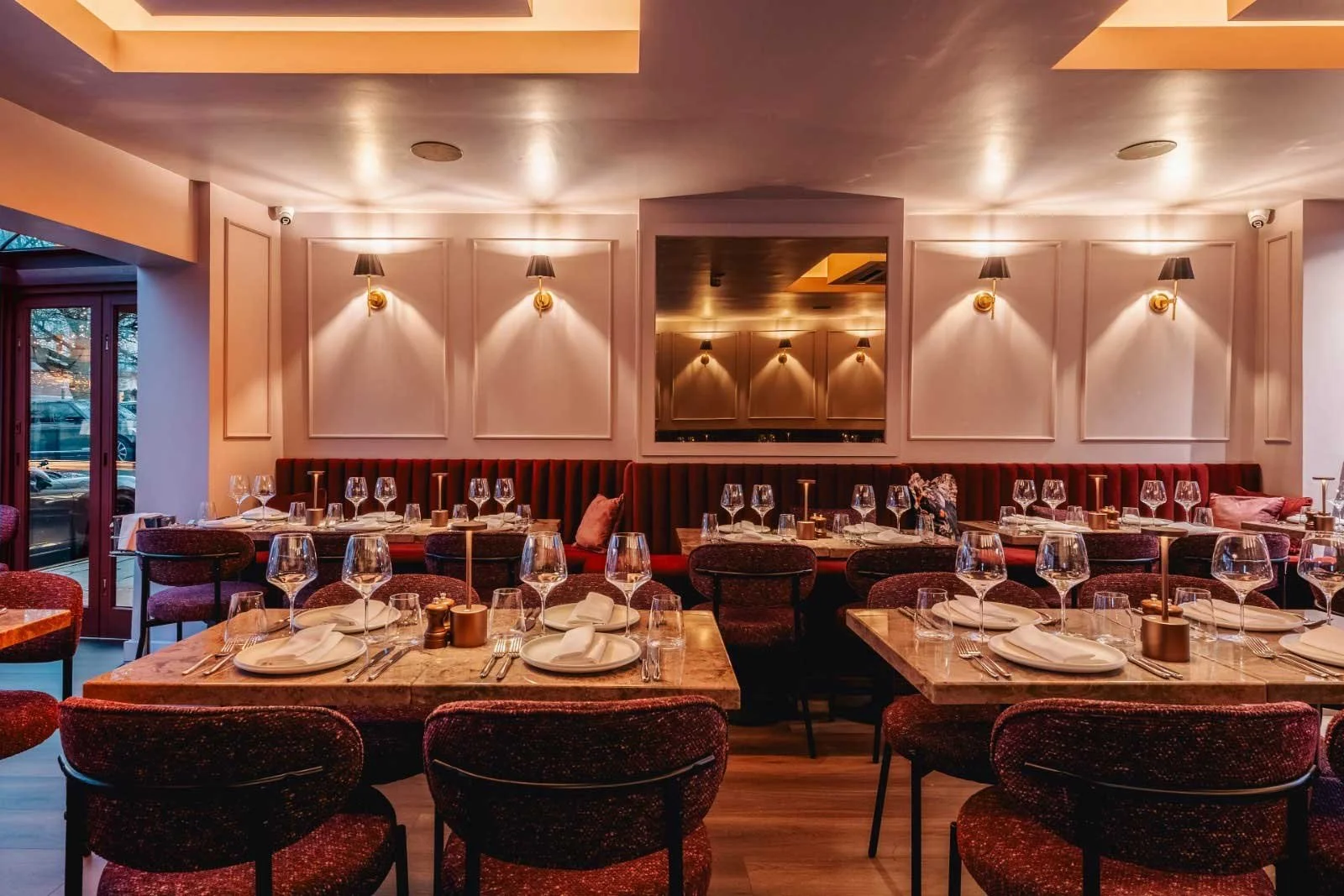 Elegant restaurant with neatly set tables, wine glasses, white napkins, and gold candlesticks, with upholstered chairs and a padded red banquette along the wall, softened lighting, and decorative wall panels.