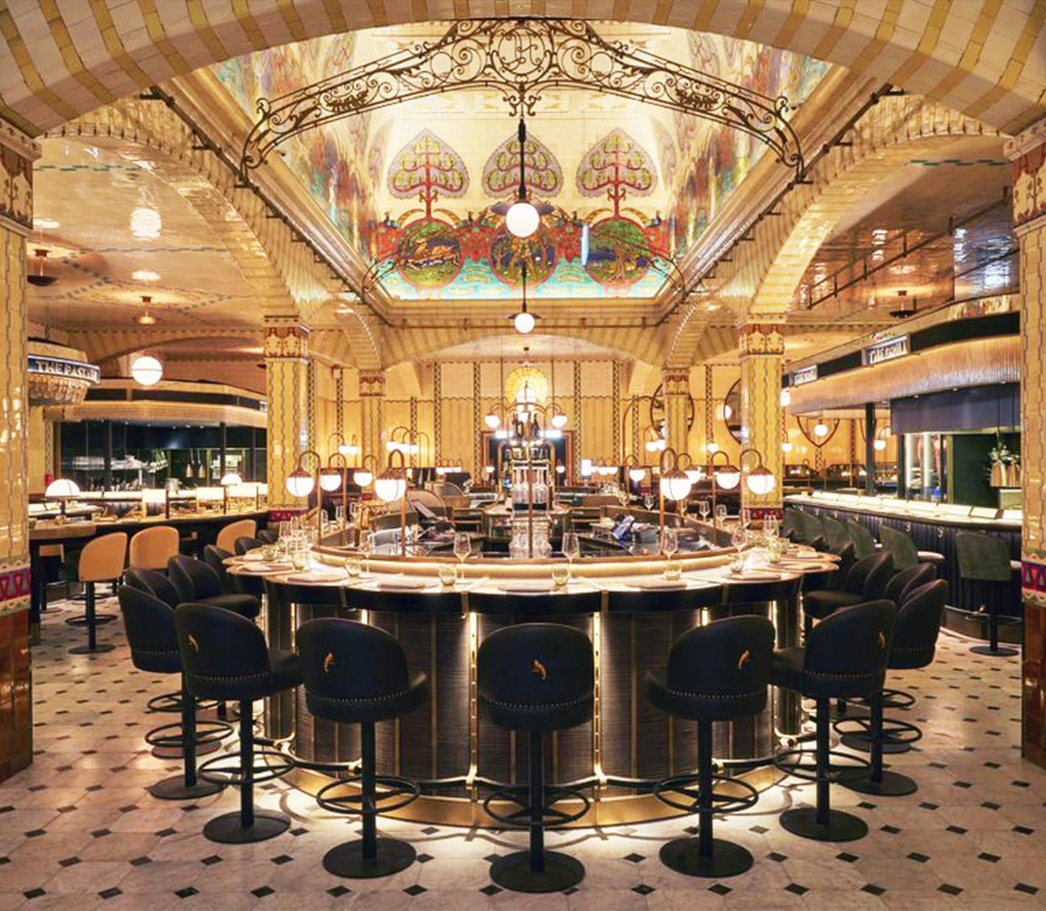 Elegant restaurant with a circular bar in the center, black chairs around it, warm lighting, ornate ceiling with stained glass artwork, and marble flooring.