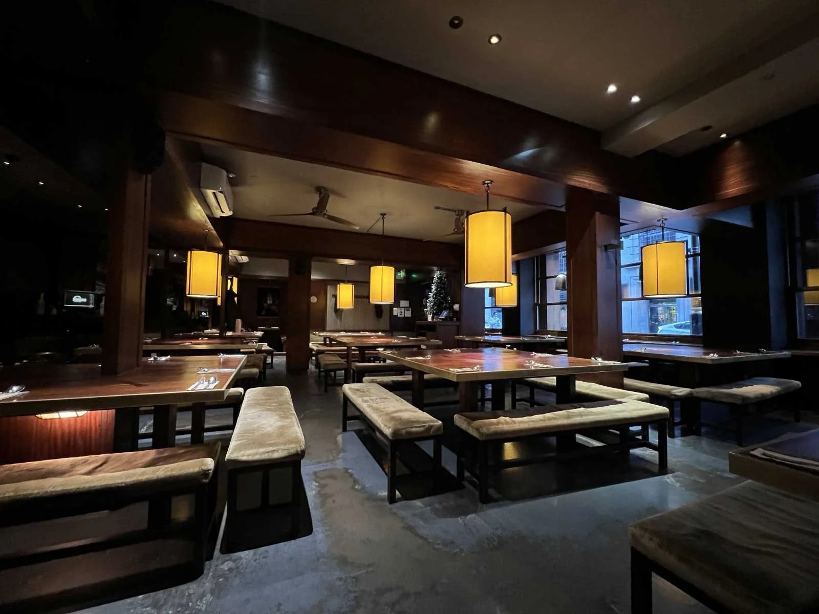 An empty restaurant interior with wooden tables, cushioned benches, large hanging yellow lamps, and large windows showing city buildings outside.