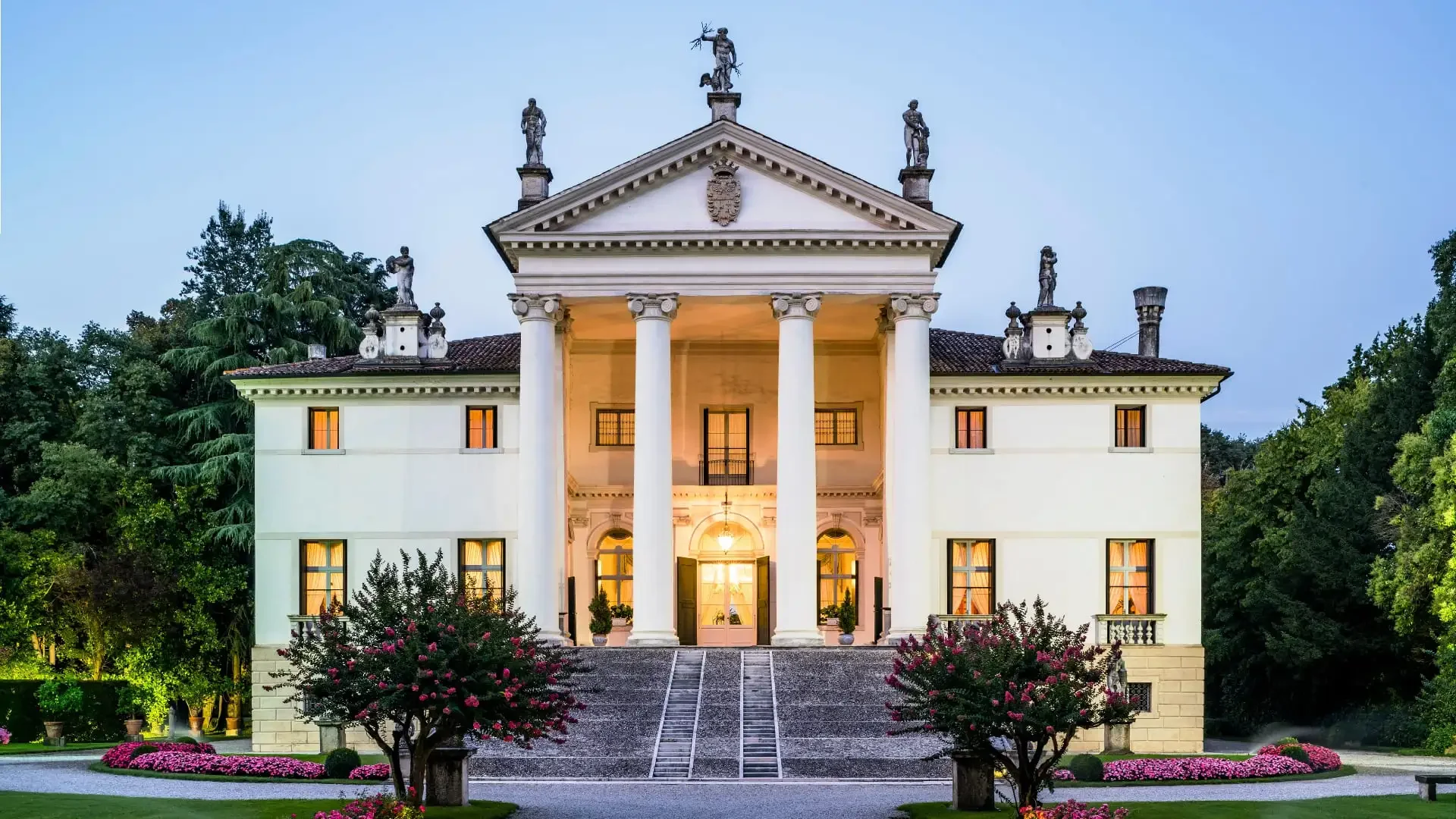 A grand white mansion with a classical architectural style featuring tall columns at the entrance, surrounded by lush landscaping and illuminated windows. Statues adorn the roofline, and a driveway with steps leads up to the front door.