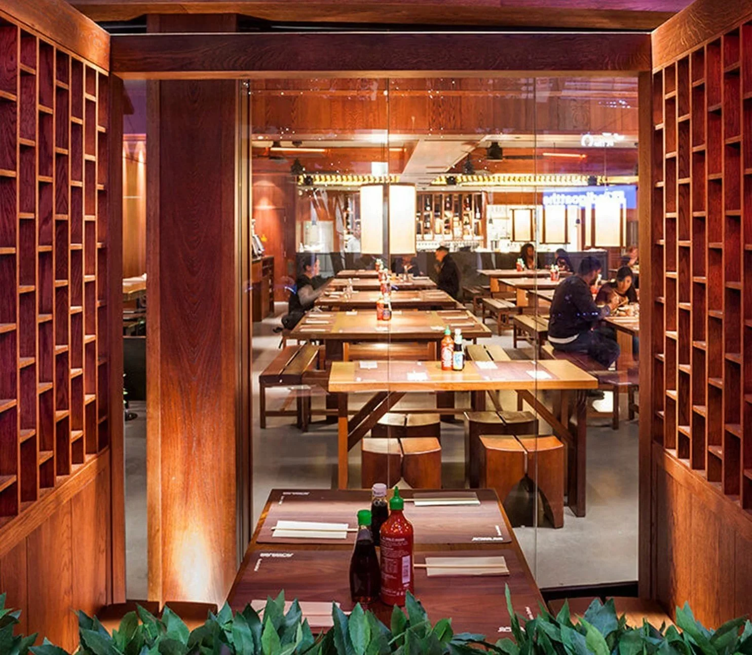 View of a restaurant interior through a window with wooden lattice framing. Inside, there are several wooden tables with condiments, and a few customers seated and dining.