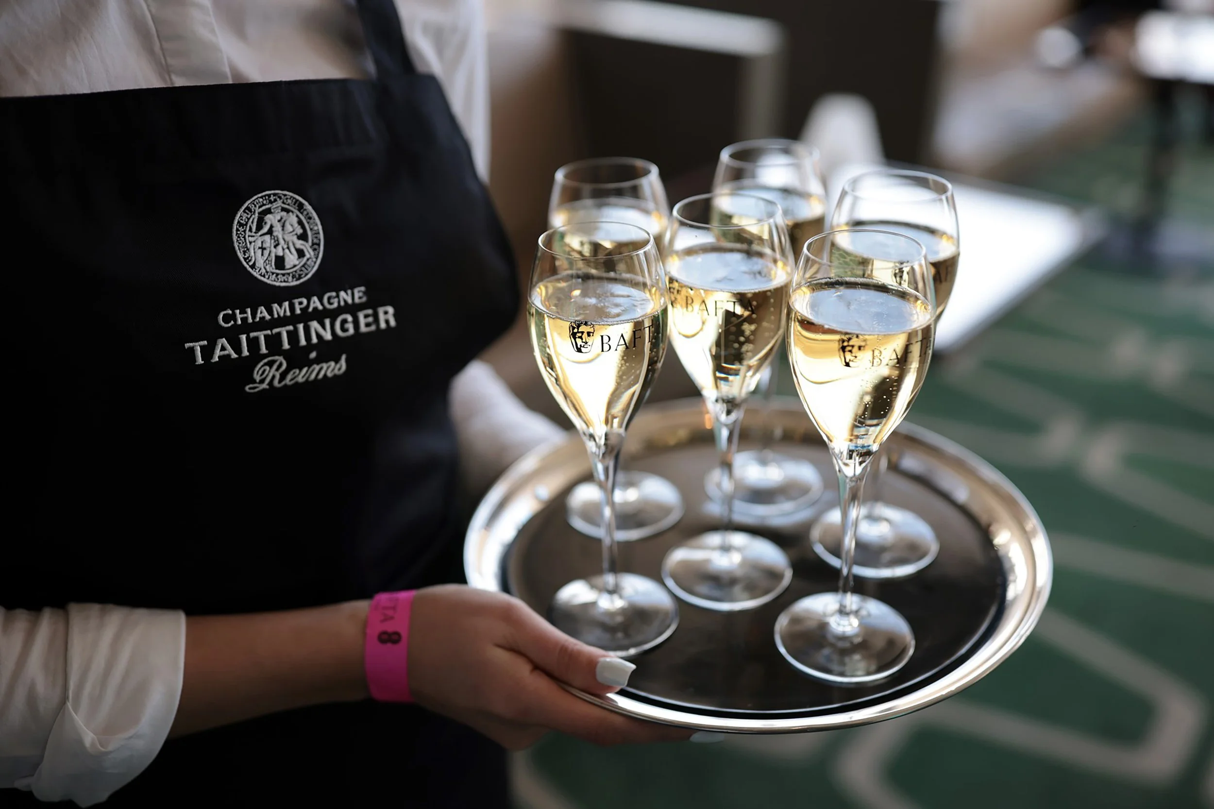 A person holding a tray with multiple glasses of champagne with a logo that says 'BAF' on each glass, and a black apron with a white logo reading 'Champagne Taittinger Reims'.