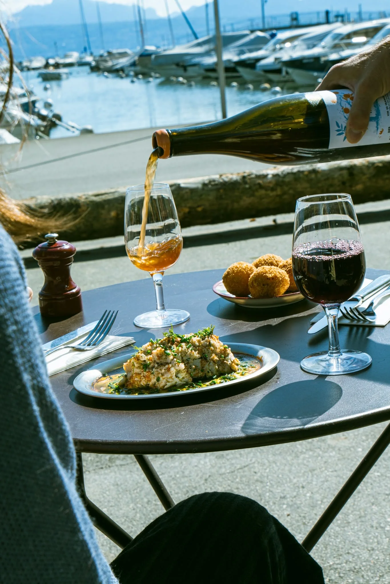 Une scène de repas en extérieur près d'un port avec des bateaux, comprenant un plat d'œufs brouillés, du pain de croûte, deux verres de vin rouge et une bouteille de vin blanc en train d'être servie, avec une personne en arrière-plan.