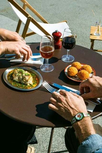 Repas en plein air avec deux personnes, un verre de vin rouge, un verre de soda, un plat de croquettes et un plat de poulet avec légumes.