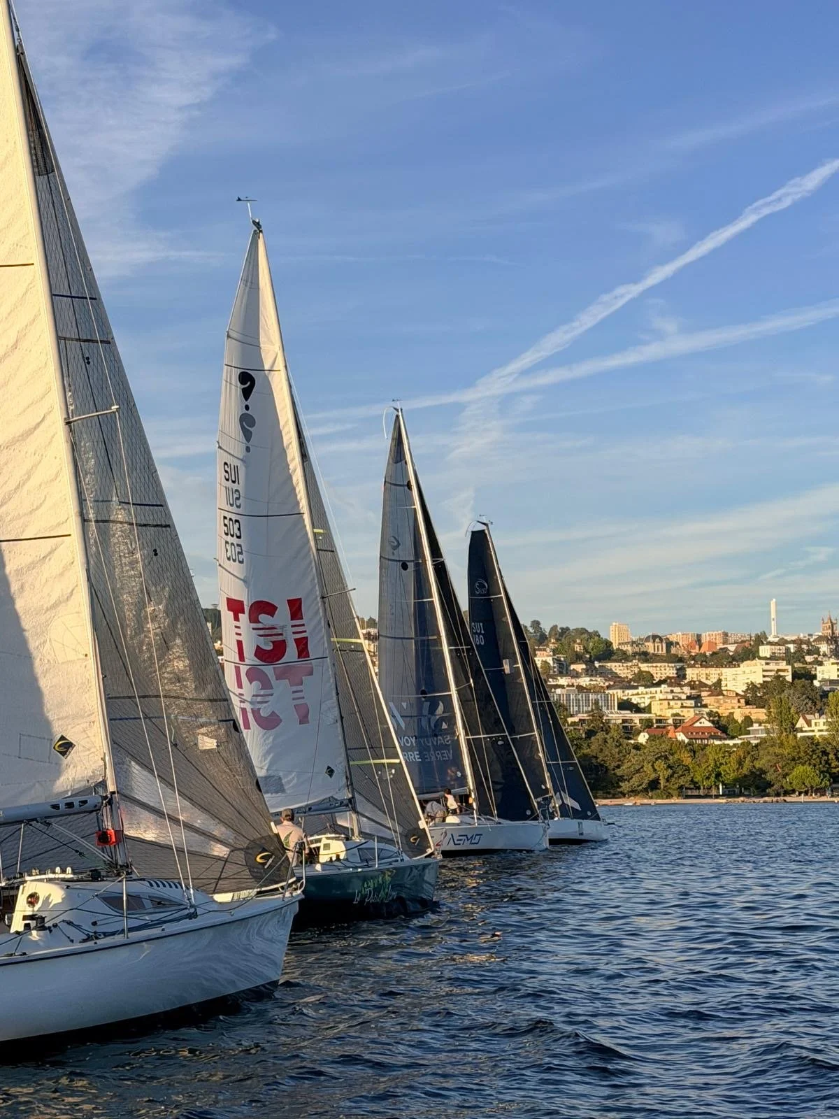 Plusieurs voiliers en course sur l'eau, avec une ville en arrière-plan sous un ciel bleu avec quelques nuages.