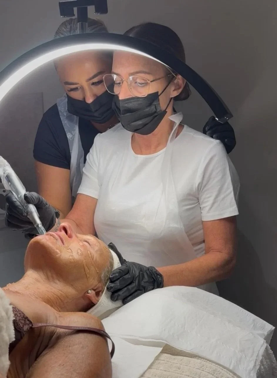 A woman wearing protective goggles and gloves performing a cosmetic treatment on a patient's legs in a clinic or spa setting.