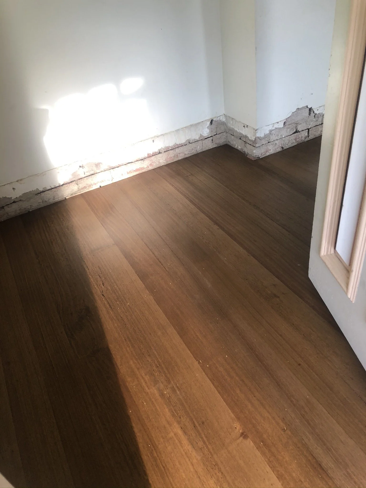 Close-up of a hardwood floor with a damaged wall corner exposing rough brick and plaster, near a white door.