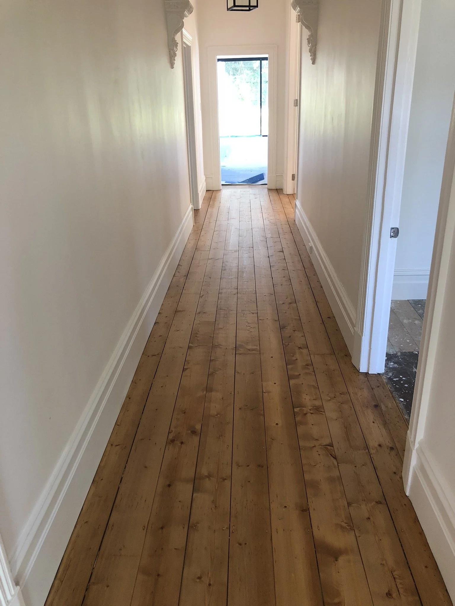 Long hallway with freshly installed wooden floorboards, cream-colored walls, white trim, and a door at the end with a glass panel leading outside.