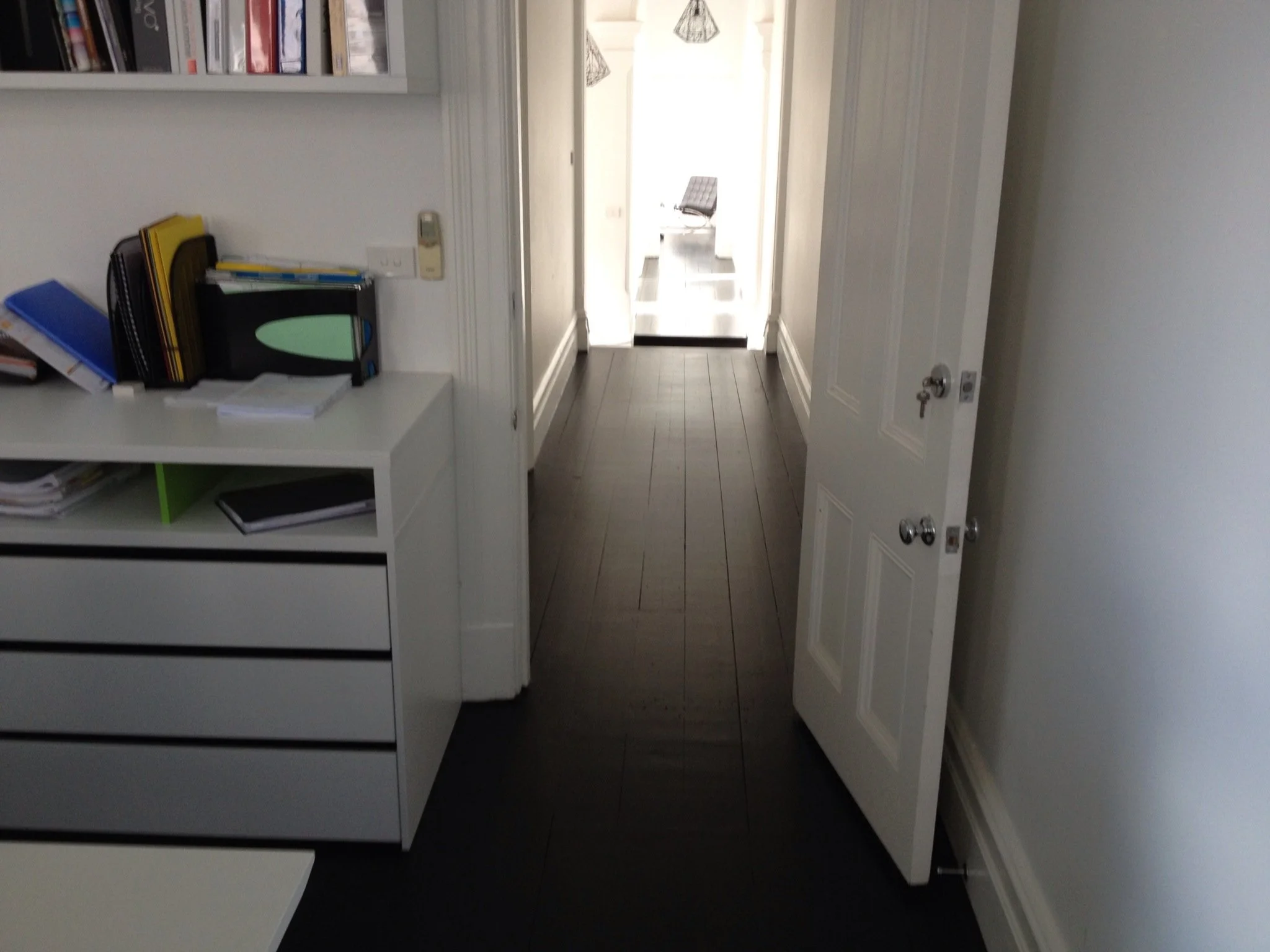 Interior view of an office hallway with a desk on the left side with various files and notebooks, white walls, dark wooden floors, and an open door leading to a brighter area with a black sofa visible in the distance.