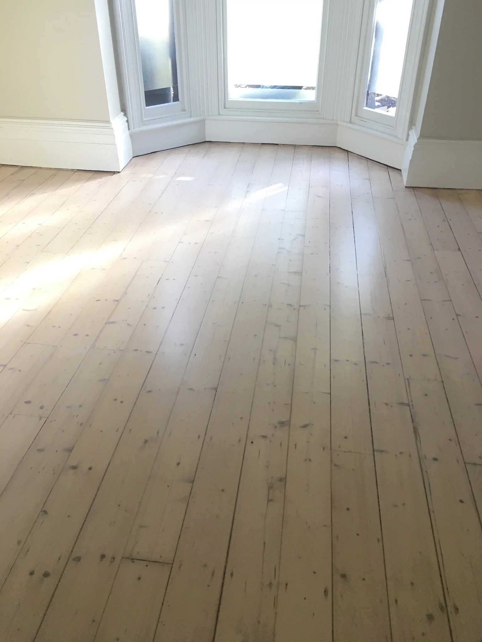 Empty room with light-colored wooden floor and large windows with white trim, letting in sunlight.