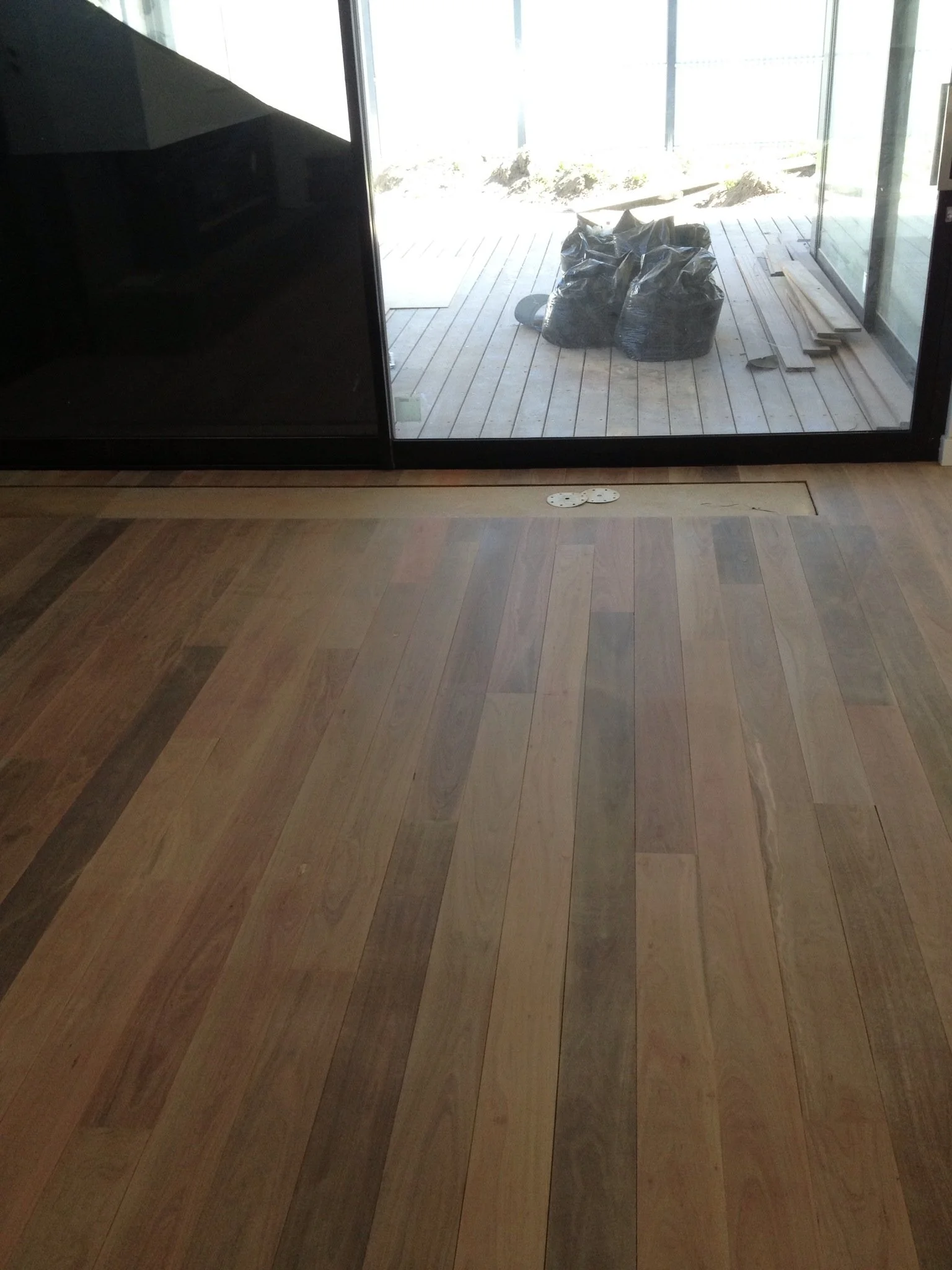 A room with wooden flooring and a glass sliding door, showing construction materials outside, including black trash bags, wooden planks, and tools.