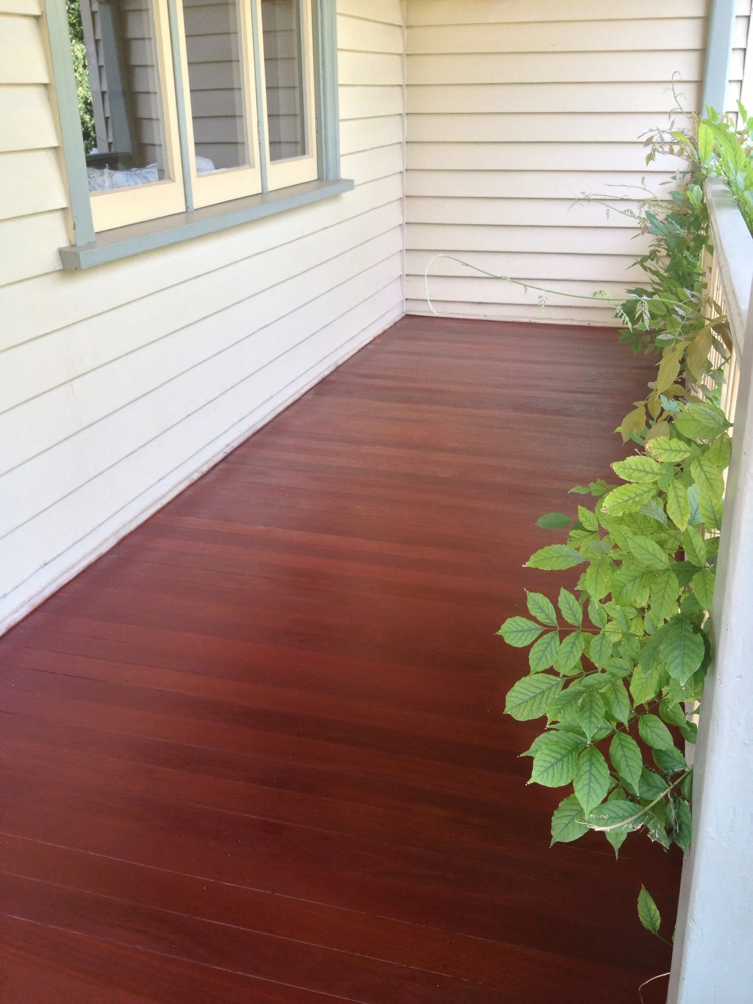 A wooden patio deck next to a house with beige siding, a window, and some green plants on the right side.