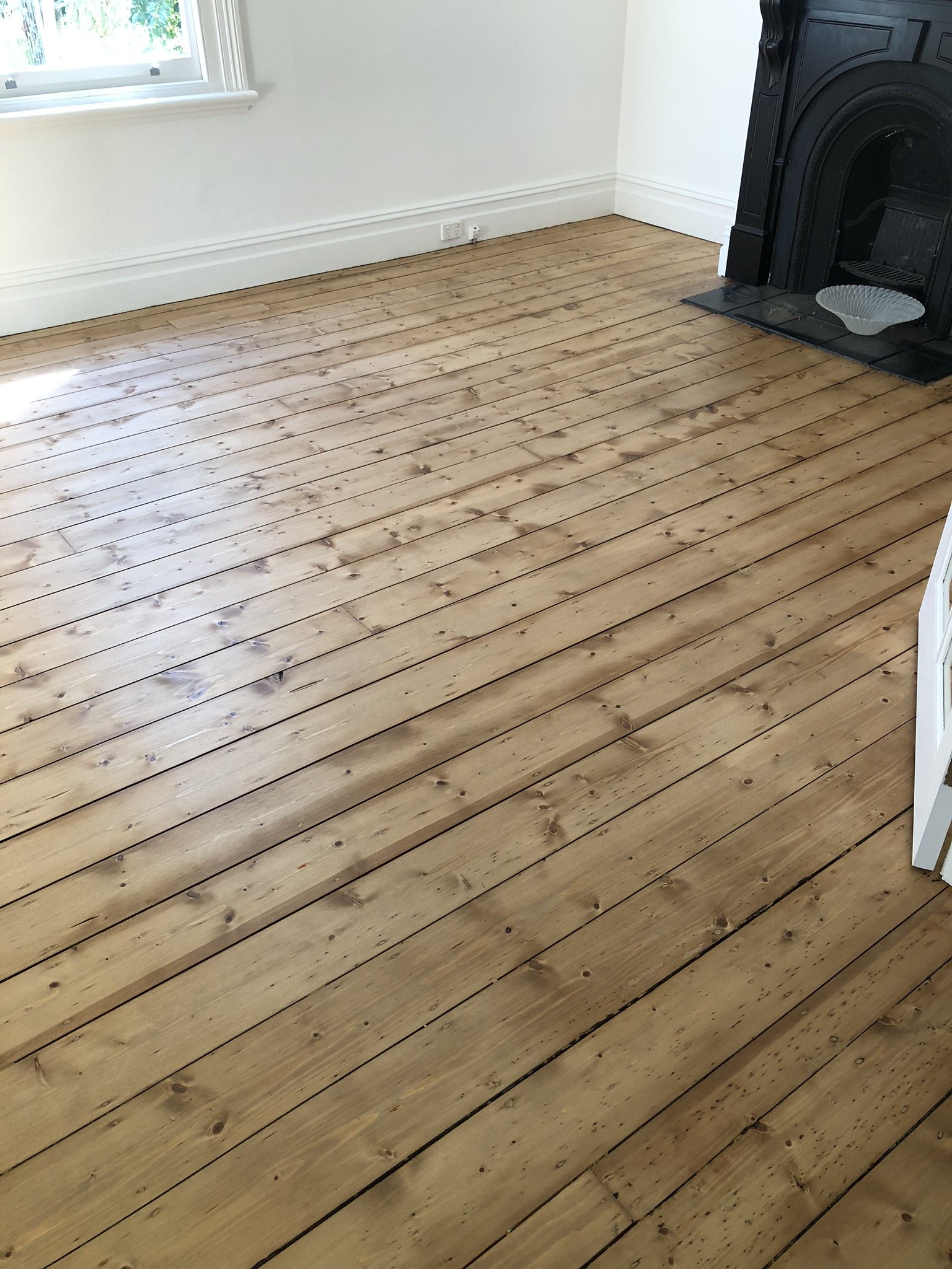 A room with newly installed wooden floorboards, a window on the left wall, and a black fireplace on the right wall. There is a small white bowl on the black hearth.