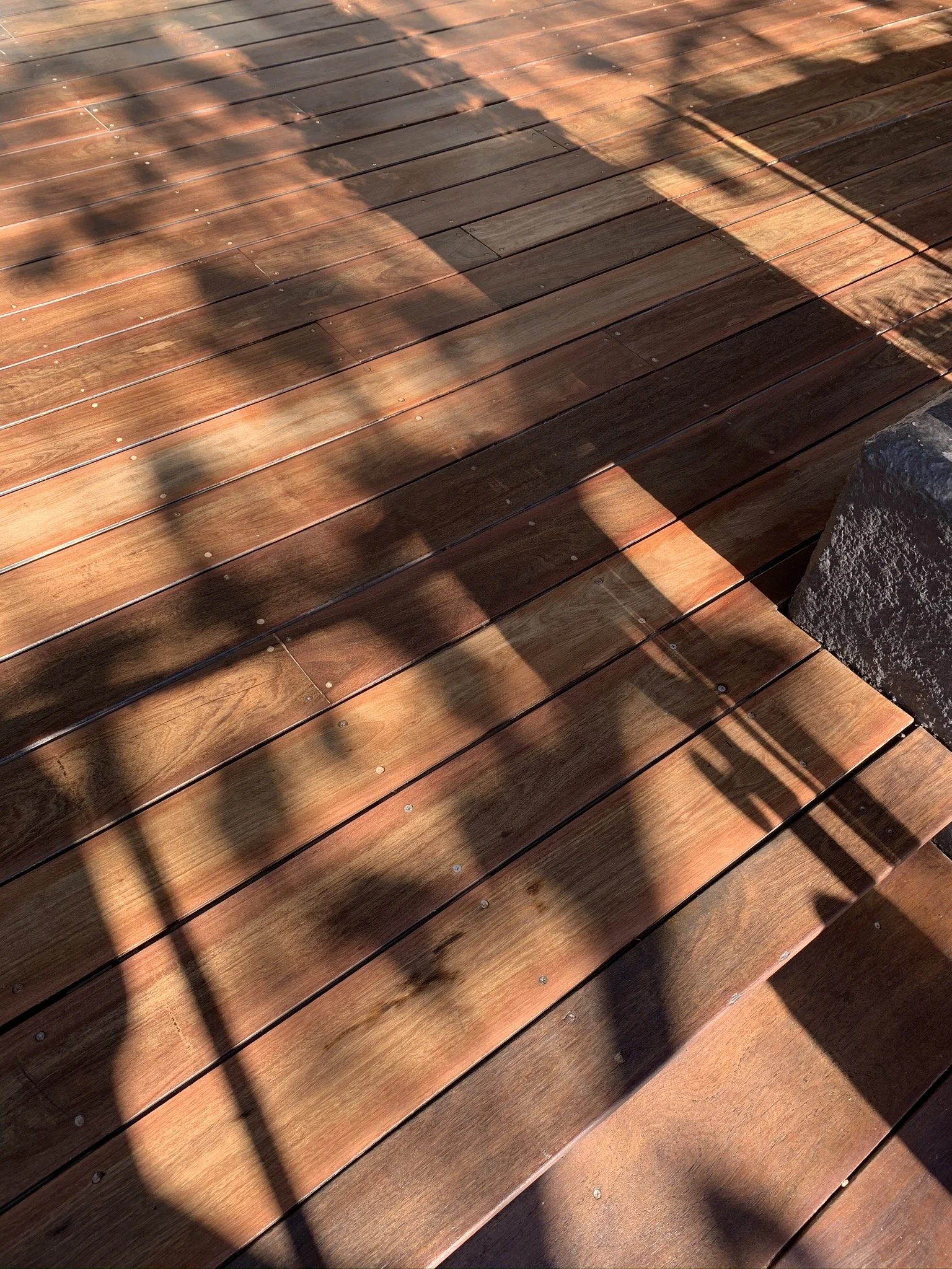 Shadow of a chair and railing cast on a polished wooden deck with a stone brick edge.