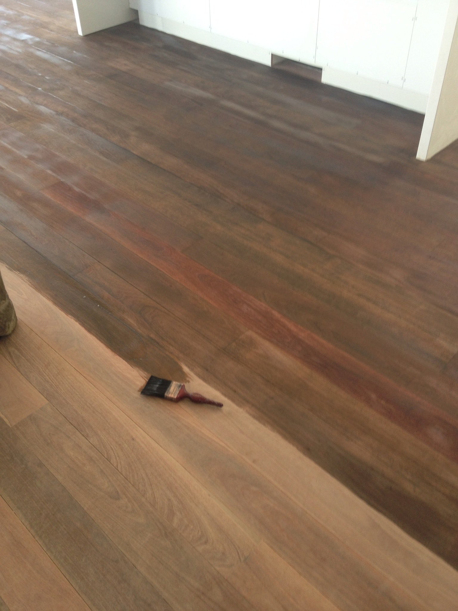 A hardwood floor being stained or finished with a brush, showing a transition from an unfinished to an finished appearance.