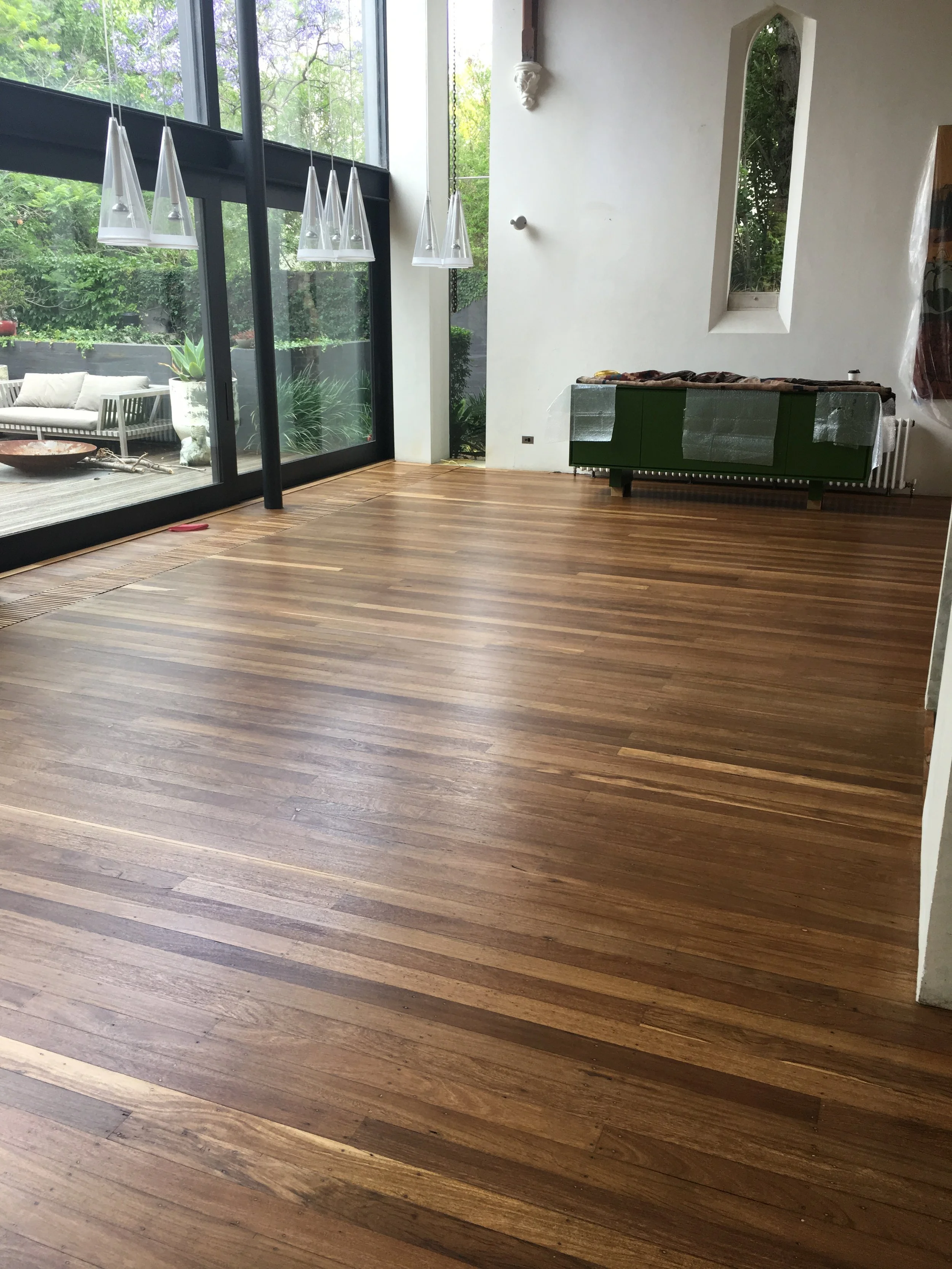 Empty room with wooden flooring, large glass sliding doors, and a small window. Some furniture is covered or unfinished, with external patio and lush greenery visible outside.