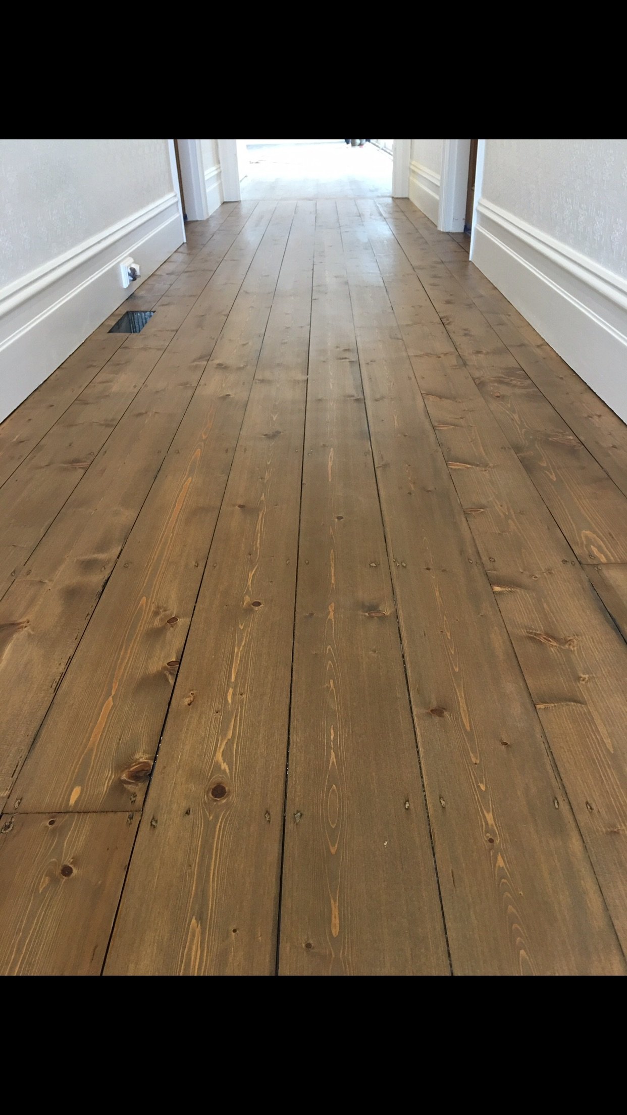 View of a hallway with light-colored wooden flooring and white baseboards. The hallway leads to a brighter area, and there is an air vent and a power outlet on the left wall.