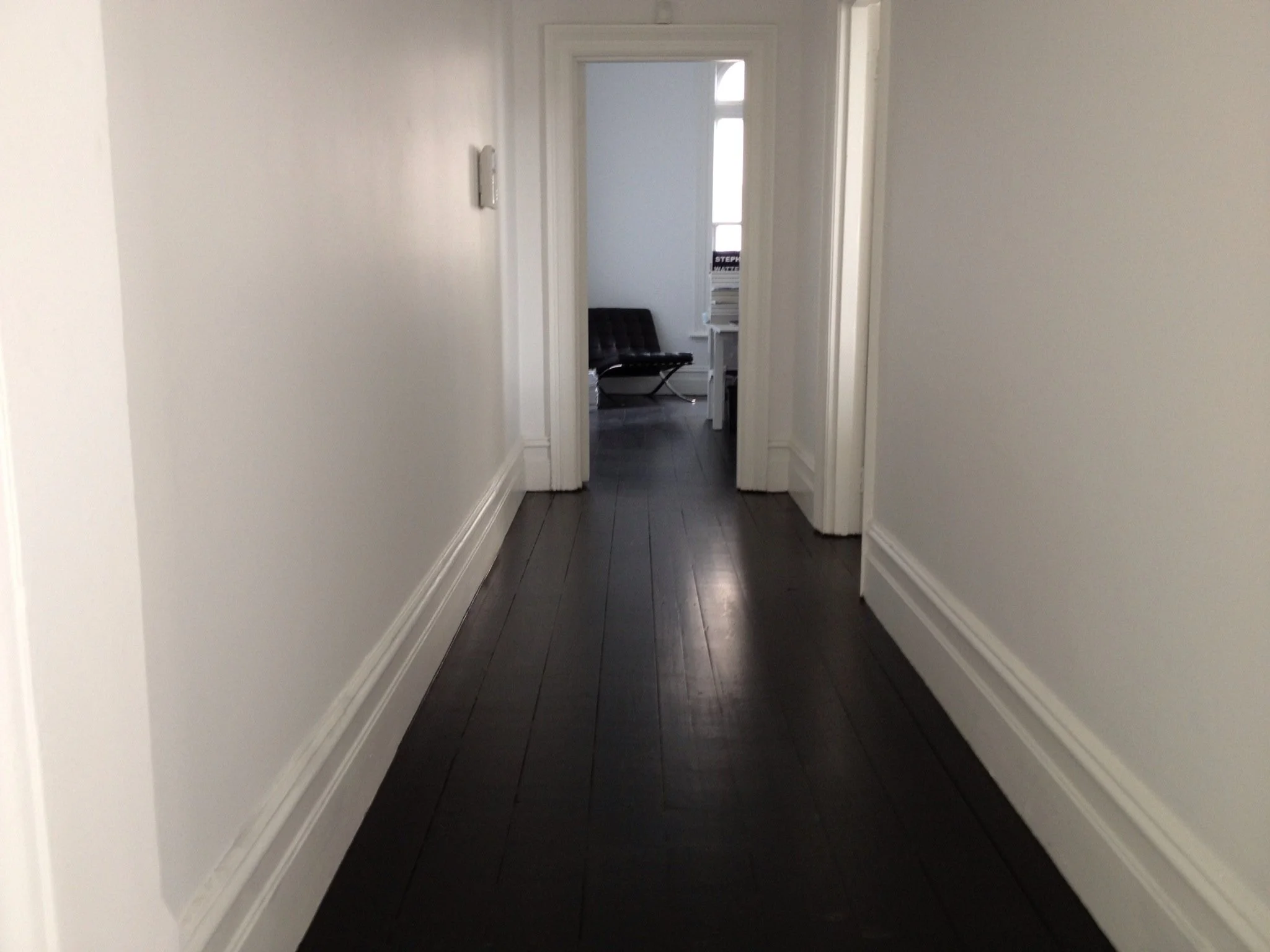 Empty hallway with white walls and dark hardwood flooring, leading to a room with black bench and stack of books visible in the background.
