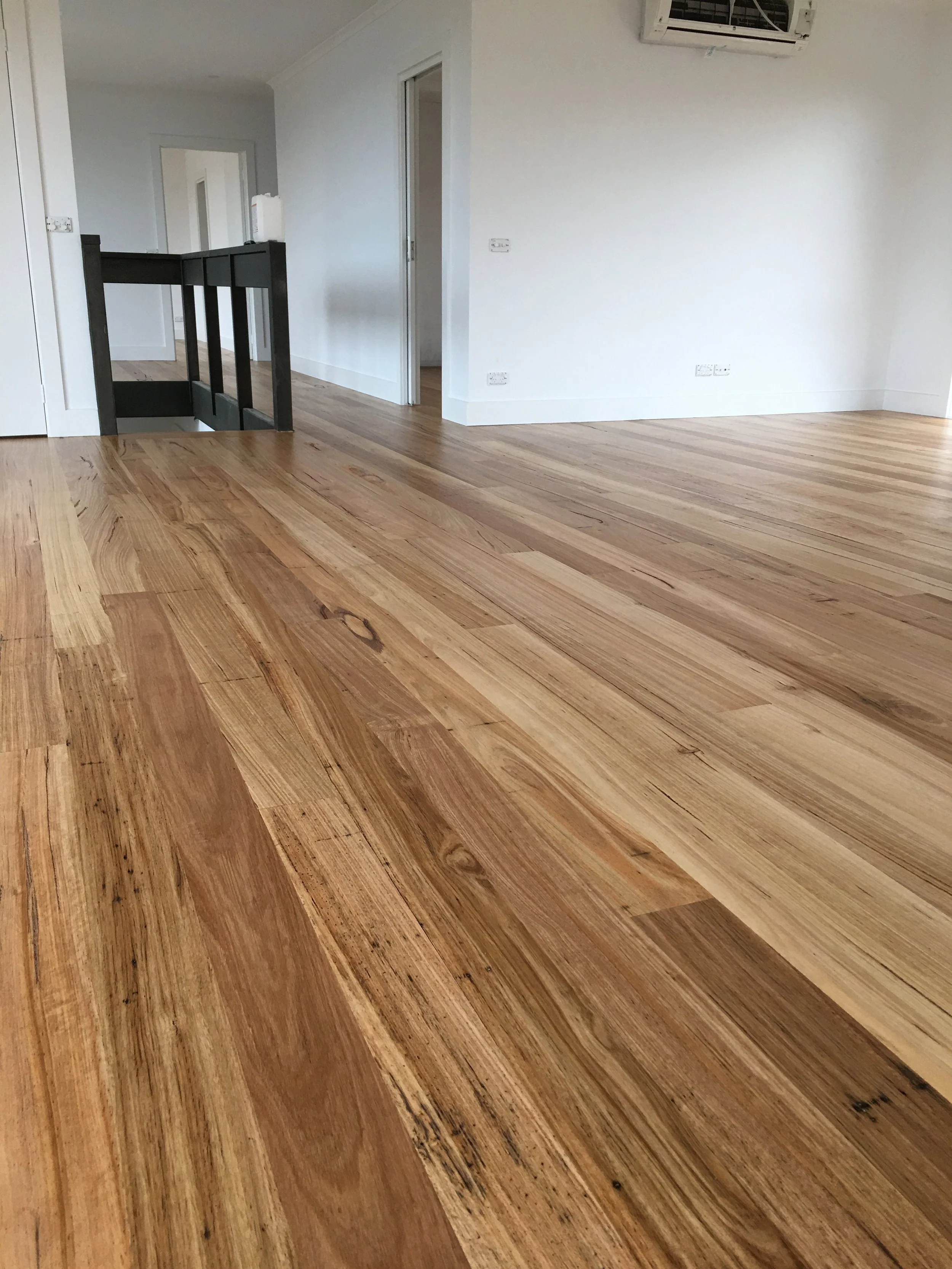 Room with polished wooden floorboards, white walls, a staircase with a black railing, and a wall-mounted air conditioning unit.