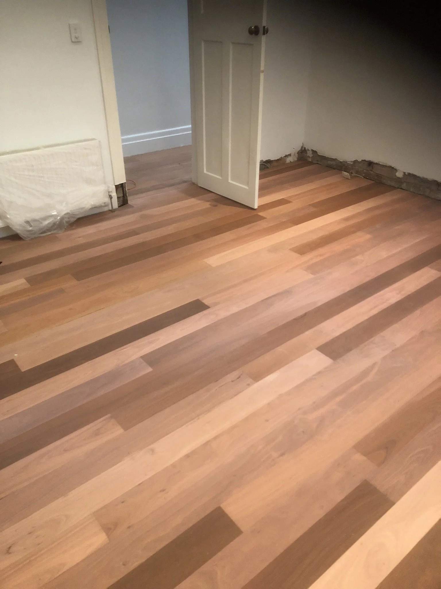 Room with new hardwood floor, open door, partially exposed wall, and a radiator covered with plastic