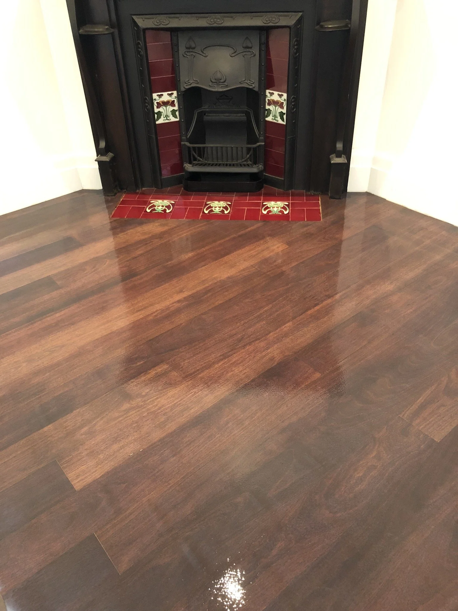 A fireplace with a black metal insert surrounded by decorative red and floral tiles, with a hearth of red tiles featuring floral motifs. The floor in front of the fireplace is polished hardwood.
