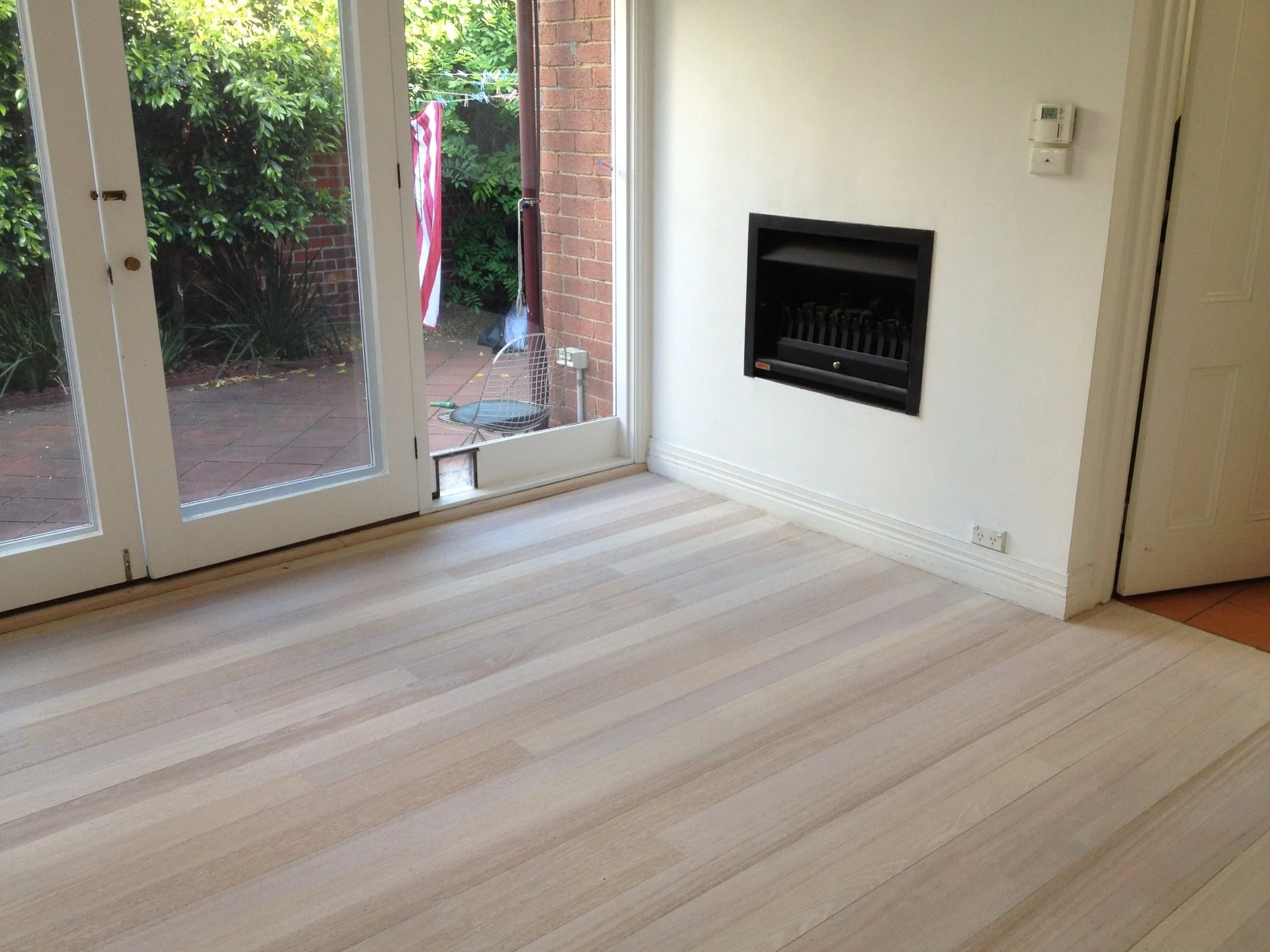 Empty room with light-colored wooden flooring, sliding glass door leading to a small patio with garden plants, a brick wall, and a white wall with a black fireplace and thermostat.