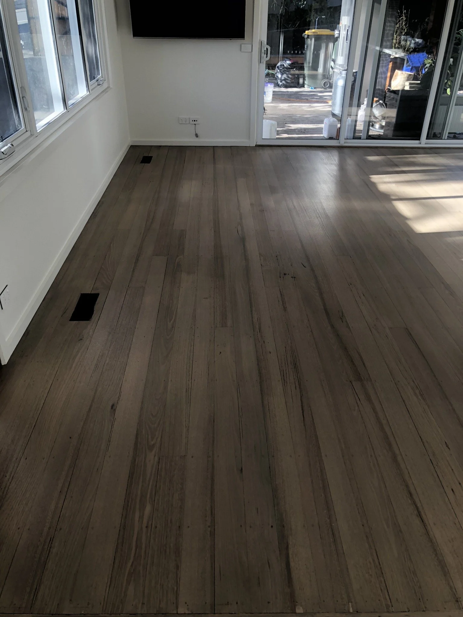 Empty room with hardwood flooring, large windows on the left, and sliding glass doors at the back leading outside, with some outdoor items visible.