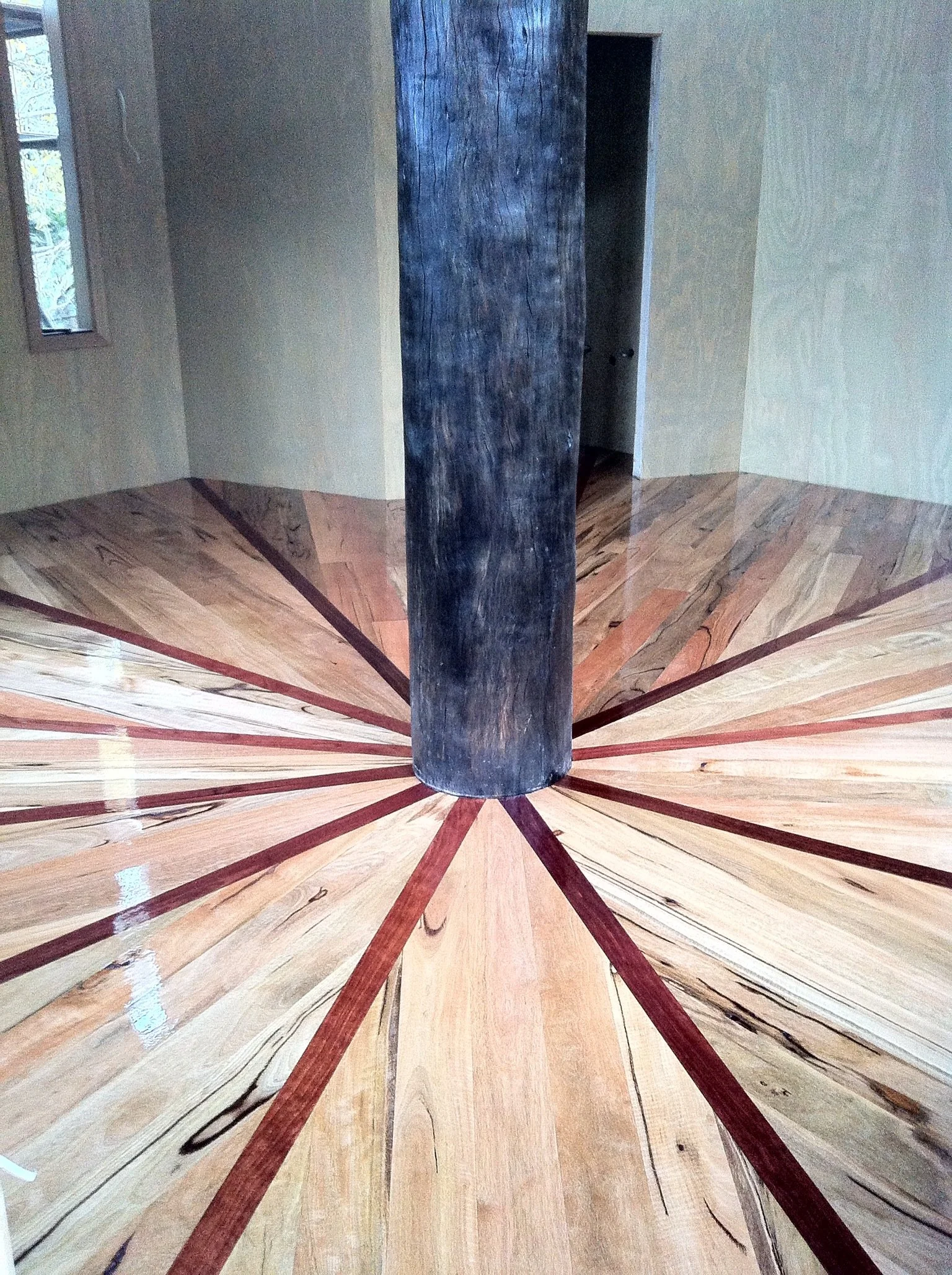 A wooden room with a central dark wooden beam and a floor pattern radiating outward from the beam with various shades of wood.