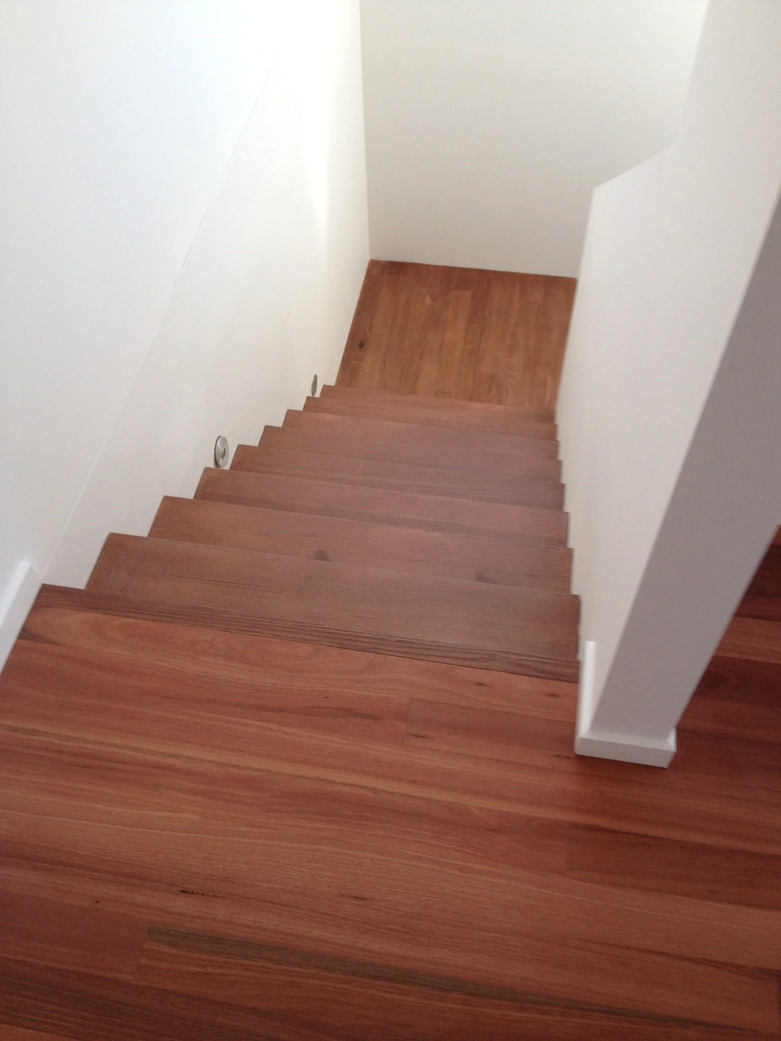 A staircase with wooden steps and white walls, leading down to a small landing with matching wood flooring.