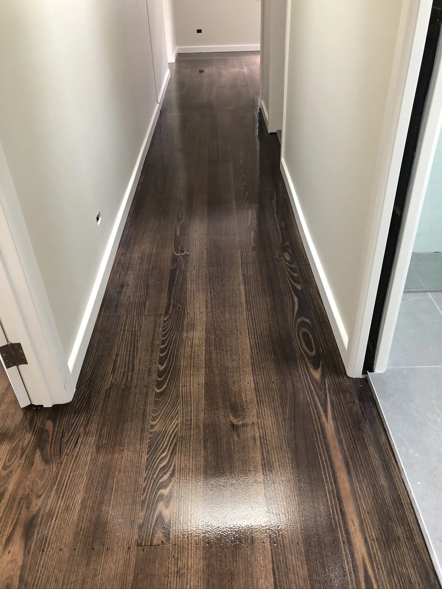 A hallway with newly finished dark wood flooring, white baseboards, and off-white walls, leading to a room at the end.