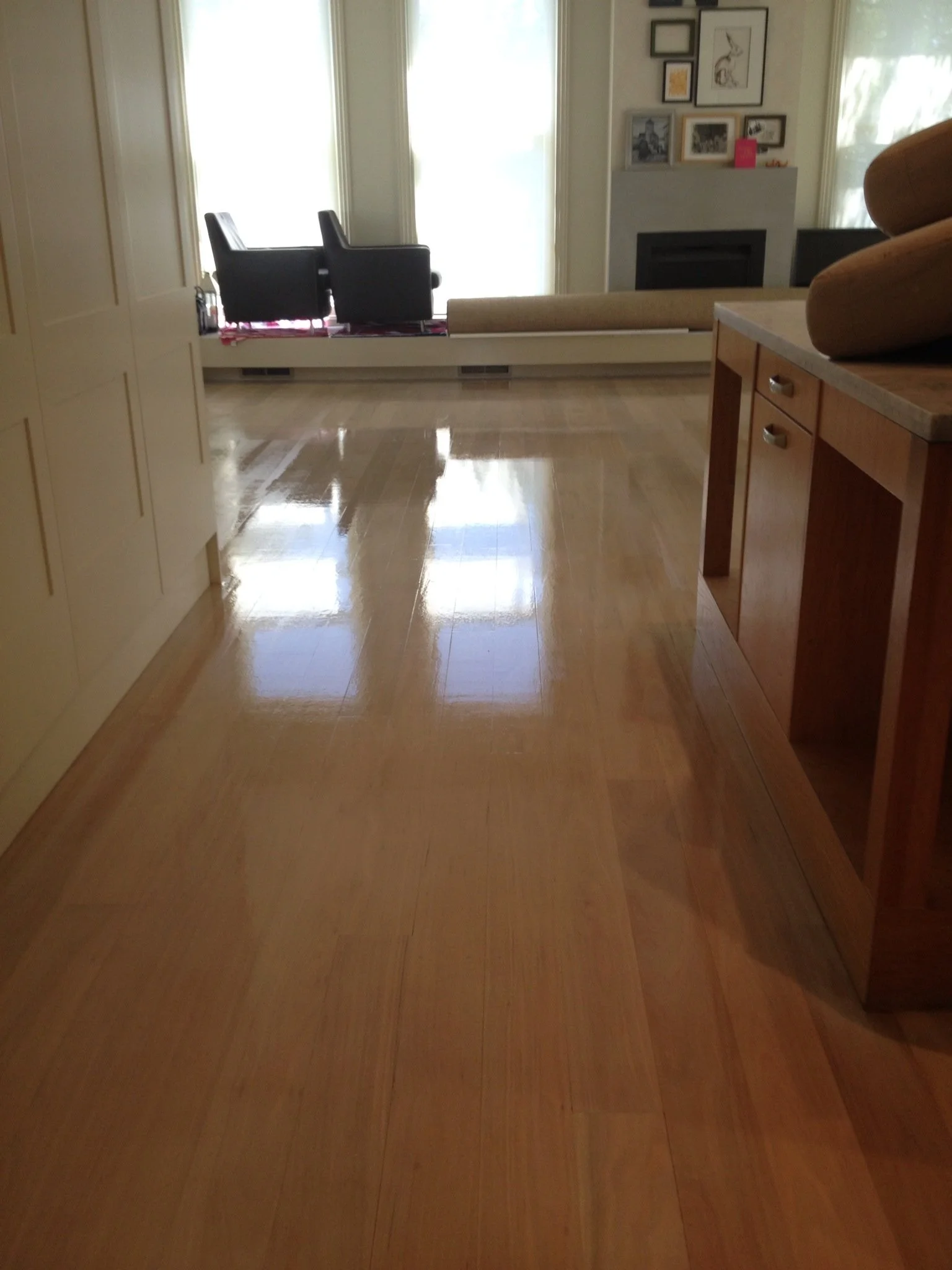 View of a shiny wooden floor in a room with large windows, a built-in window bench with two black chairs, and wall art above a fireplace.