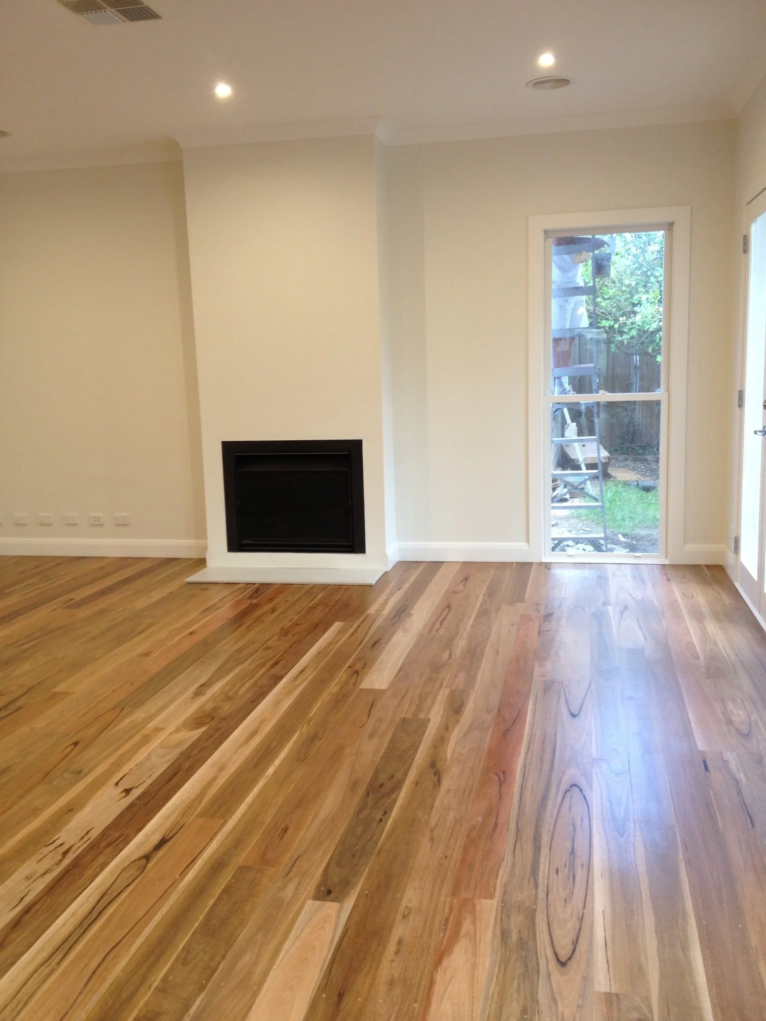Empty living room with hardwood flooring, white walls, a small fireplace, and a glass door leading outside with a ladder visible through it.