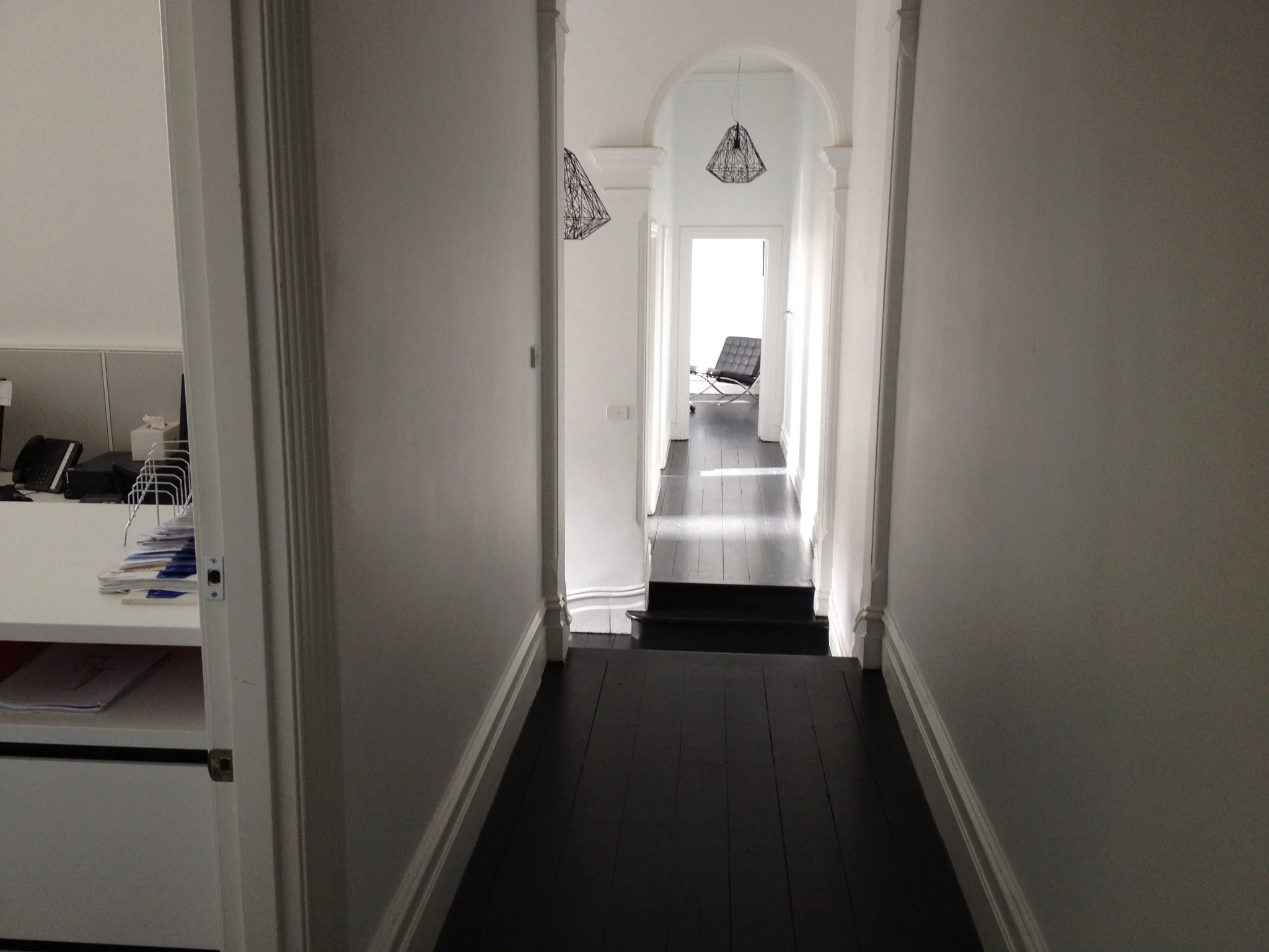 A hallway with white walls, dark wood flooring, and black pendant light fixtures, leading to a bright room with a black lounge chair near a large window.