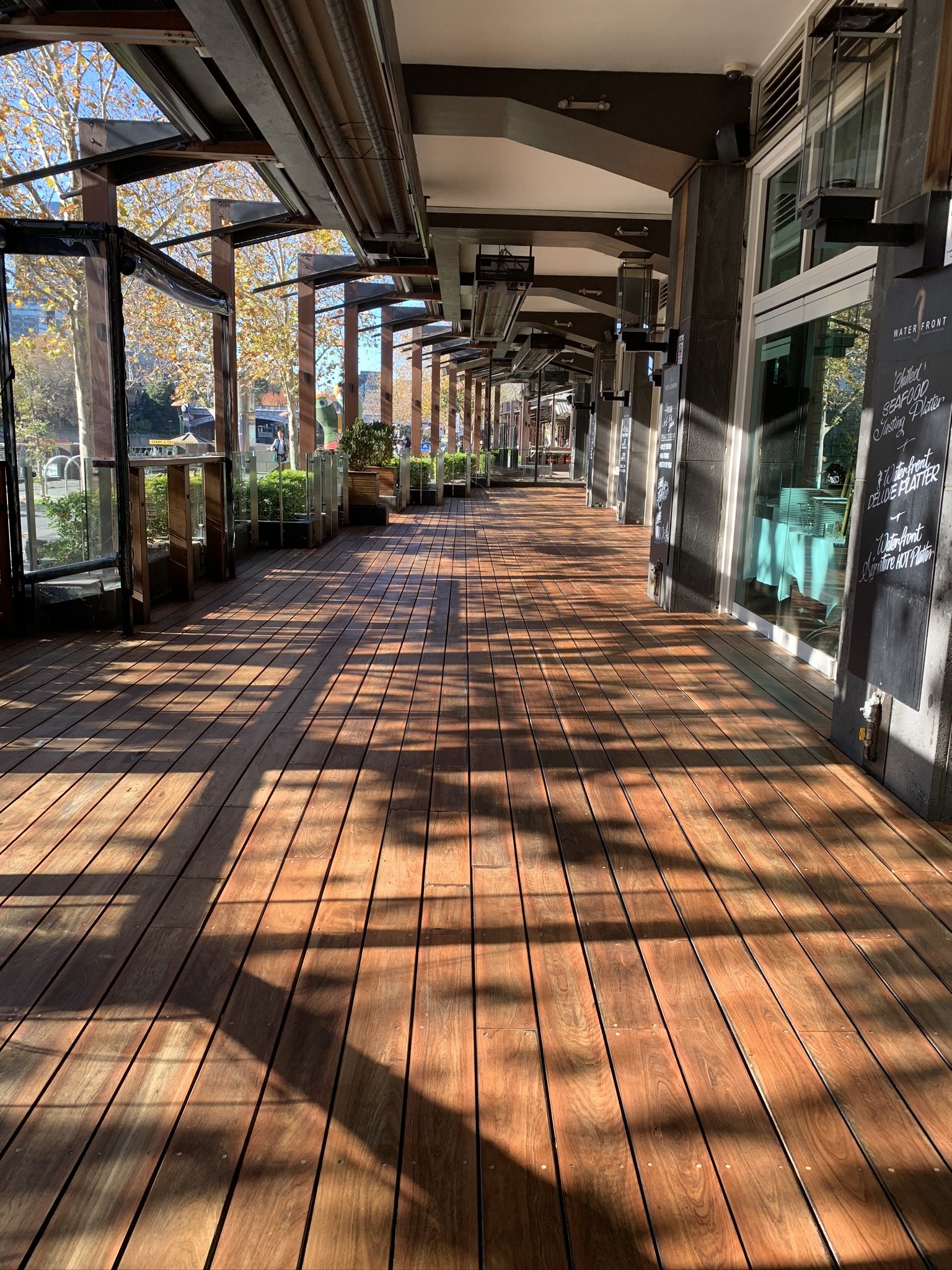 Empty outdoor patio or walkway with wooden flooring, shaded by an awning, with open windows and some potted plants along the side.