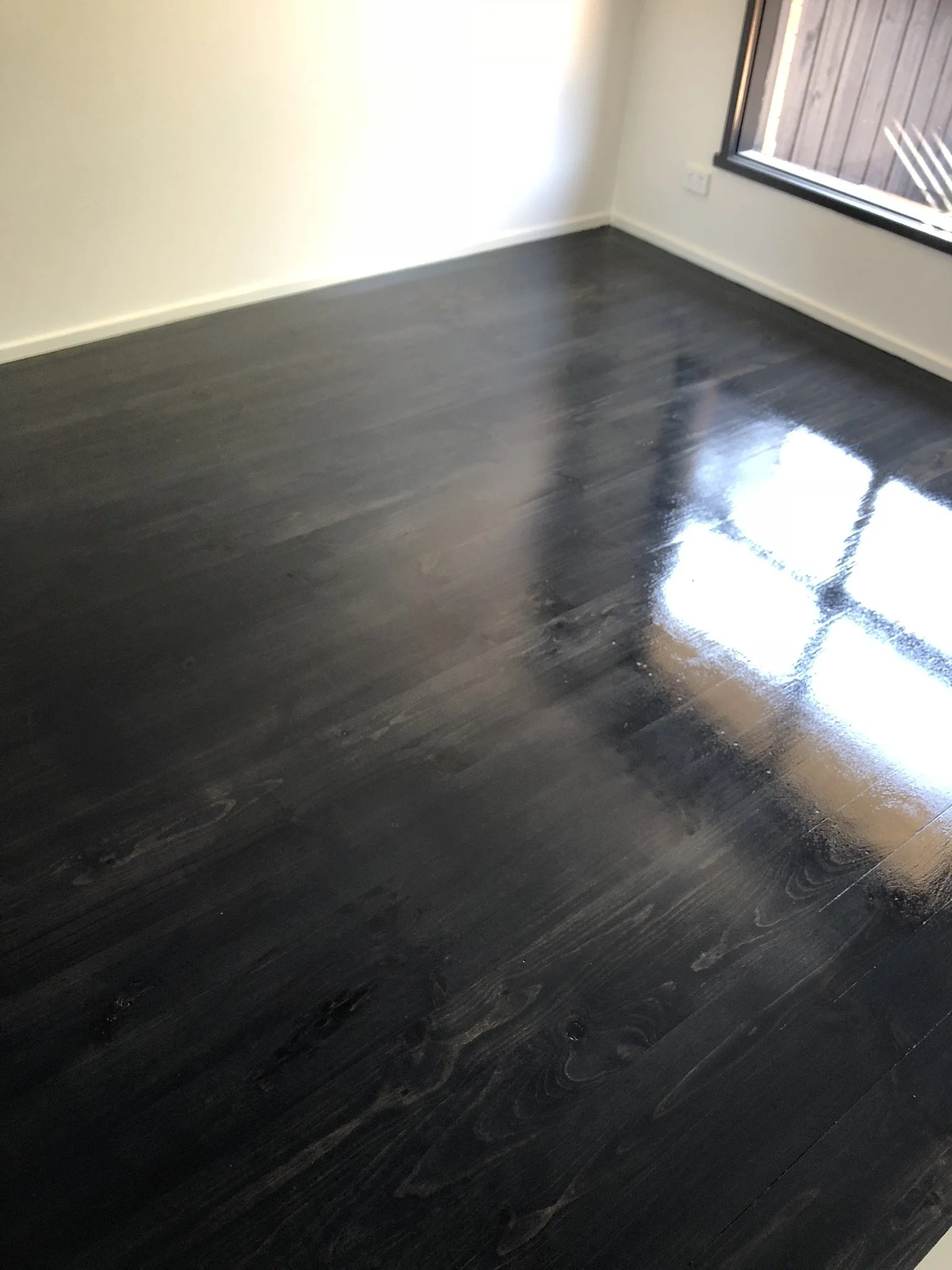 Room with dark, glossy wooden flooring and a large window showing an outside wooden fence.