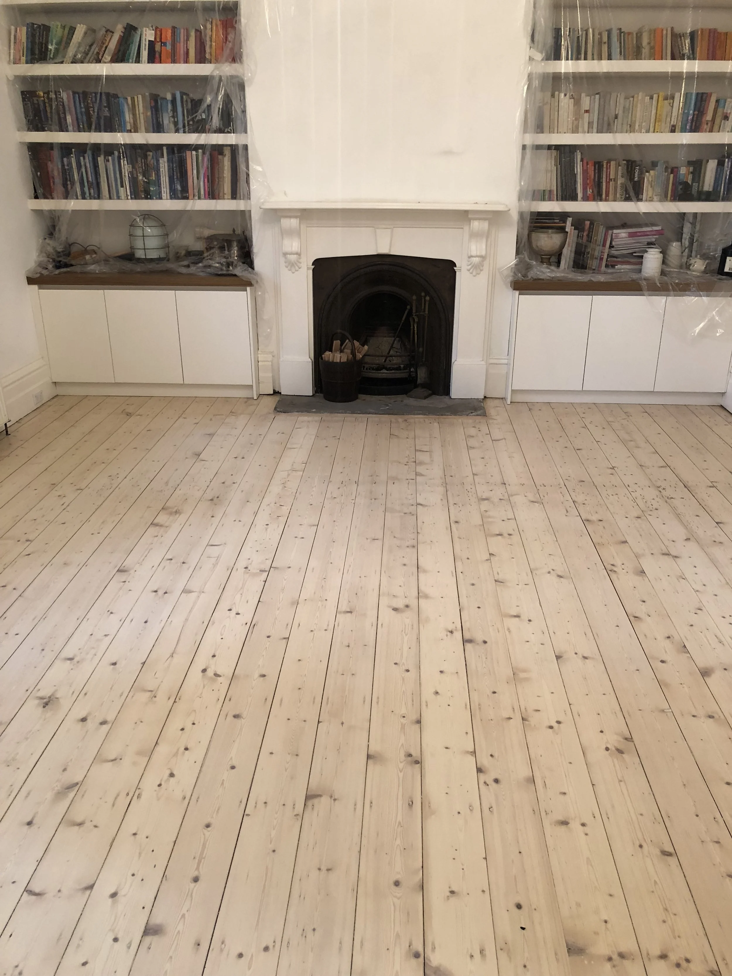 Living room with light wood floor, white fireplace with black insert, and built-in white bookshelves filled with books, covered with plastic sheets.