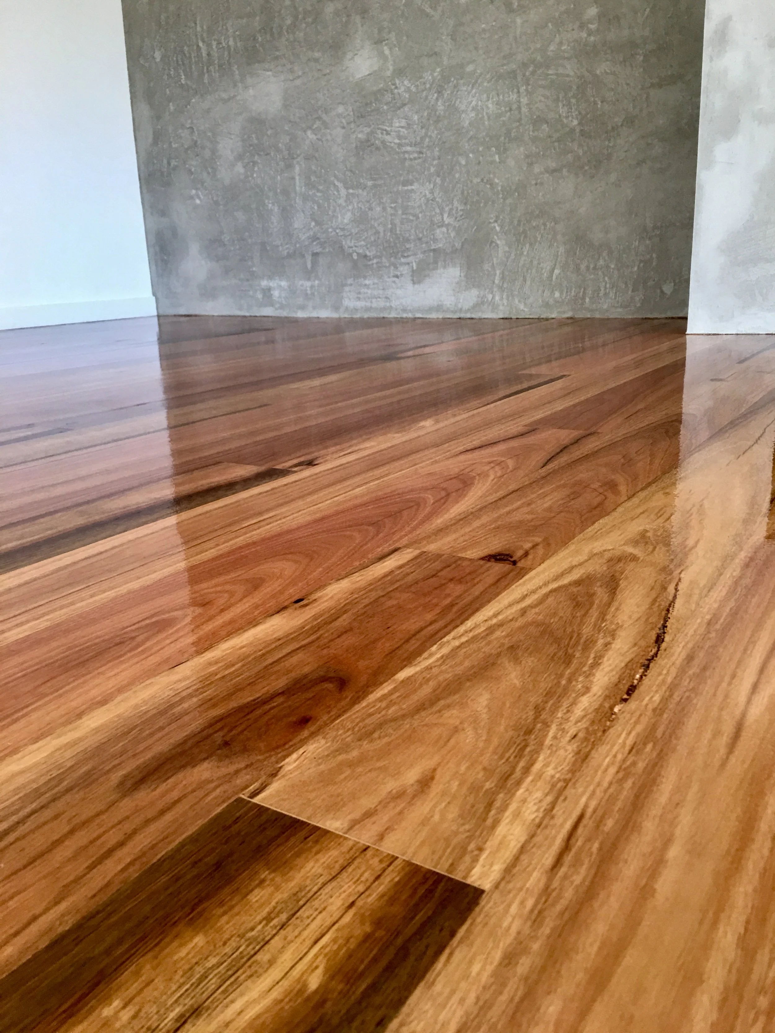 Close-up view of a polished wooden floor with a natural grain pattern and a smooth finish, with a textured wall in the background.